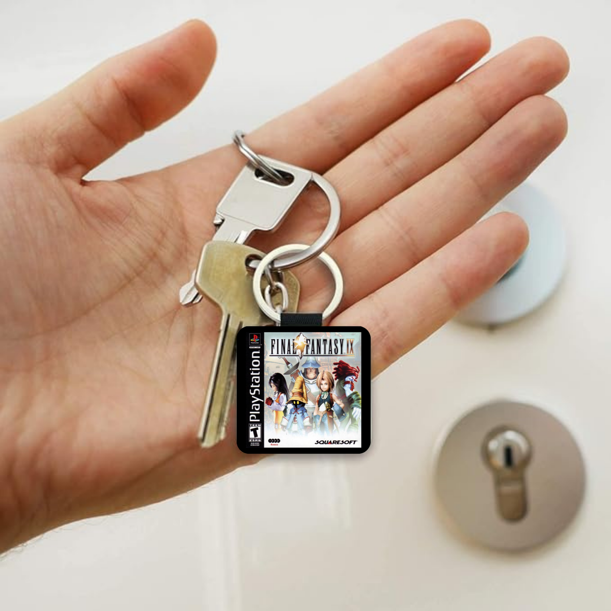 A hand holding a silver keyring with a square black keychain featuring the 'Final Fantasy IX' PlayStation game cover art, with two metal keys attached, against a light background with blurred lockpicks.