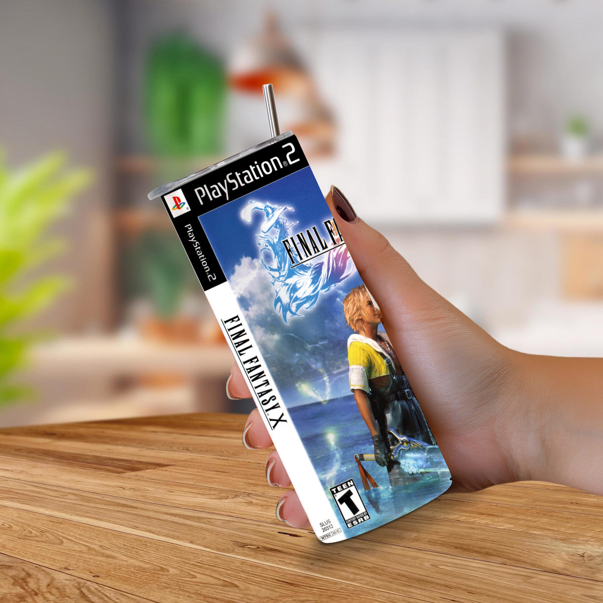 A hand holds a PlayStation 2 game 20oz tumbler with a metal straw, featuring Final Fantasy X artwork, against a blurred indoor background.