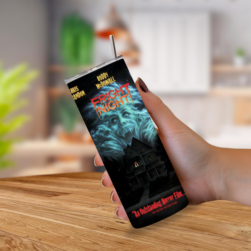 A hand holding a slim, rectangular insulated tumbler with a straw, printed with a horror movie poster design for 'Fright Night' on a wooden table with a blurred kitchen background.