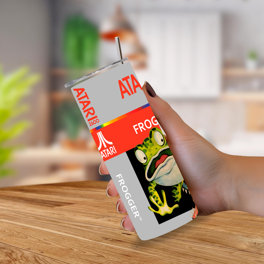 A hand holds a 20oz tumbler with a straw, printed with Atari-themed frogger graphics against a blurred indoor background with a wooden table.