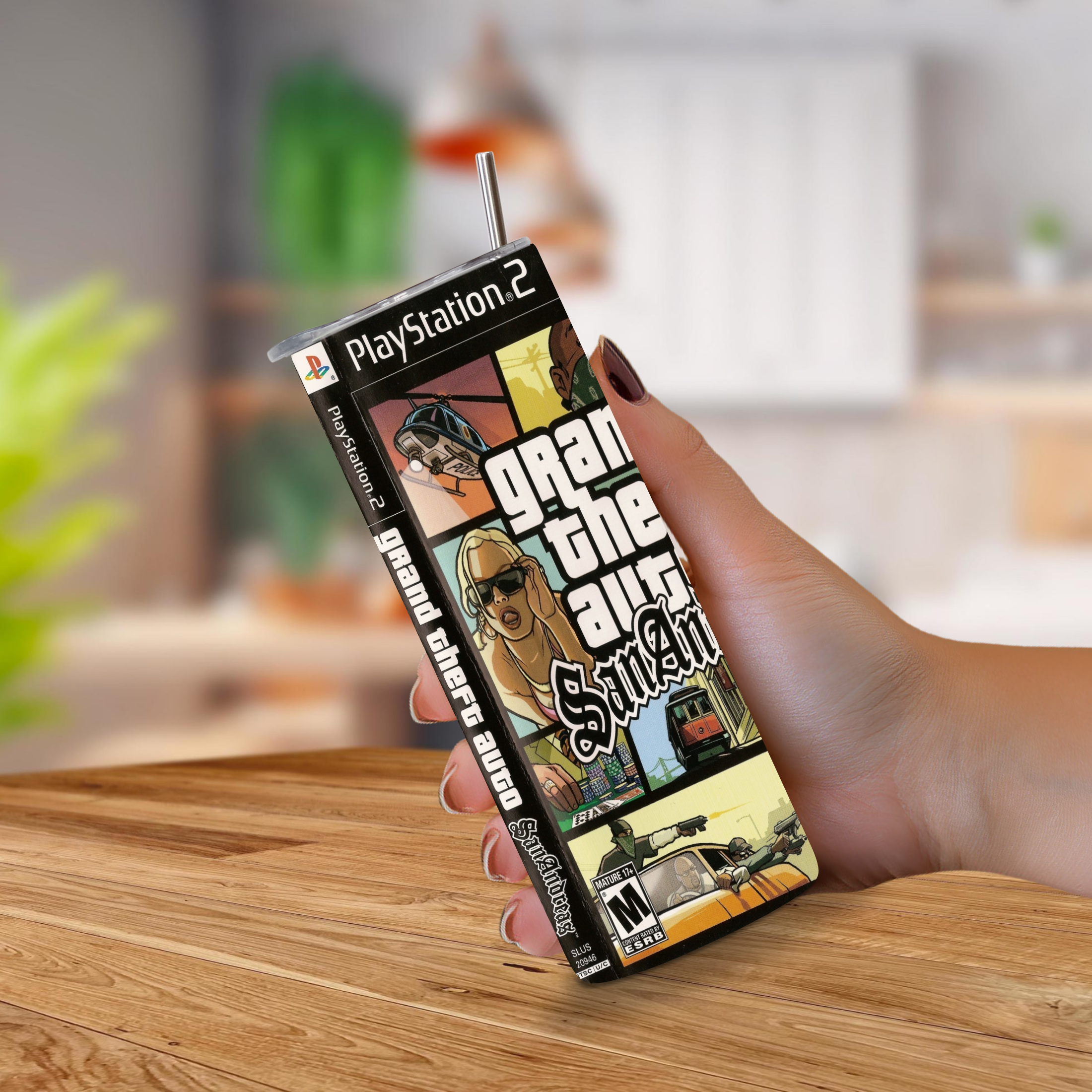 A hand holds a PlayStation 2 game cover styled as a 20oz stainless steel tumbler with a straw, featuring the Grand Theft Auto: San Andreas cover art, against a blurred indoor background.