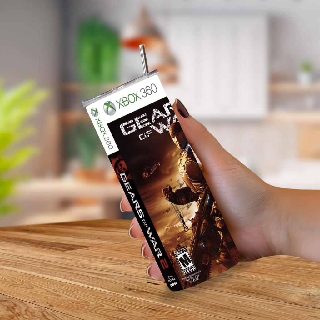 A hand holding a 20oz tumbler styled like Xbox 360 game packaging, featuring the Gears of War 2 poster art, with a straw sticking out, set against a blurred indoor background.