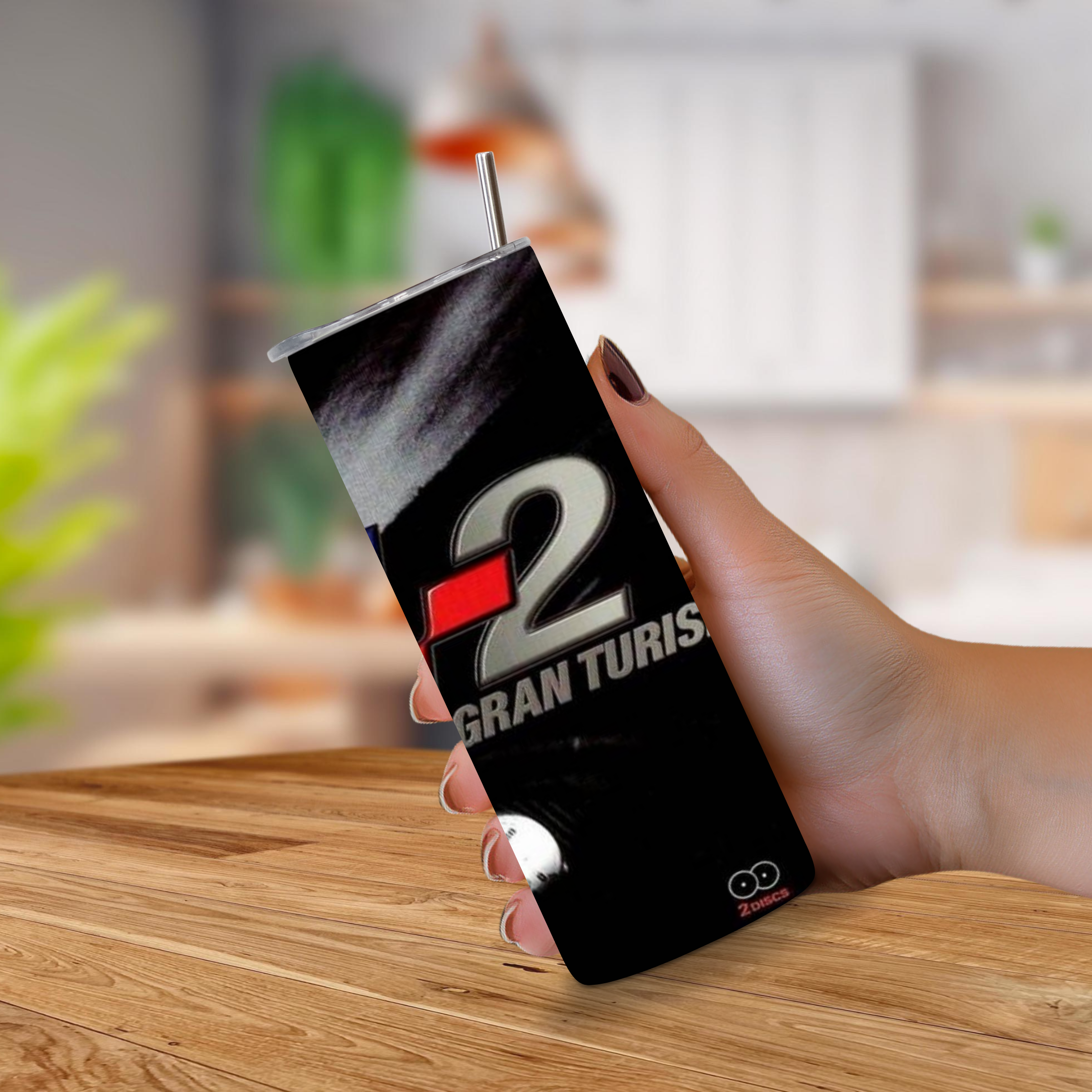 A hand holding a Gran Turismo 2 20oz stainless steel tumbler with a straw, set against a blurred kitchen background and a wooden table.
