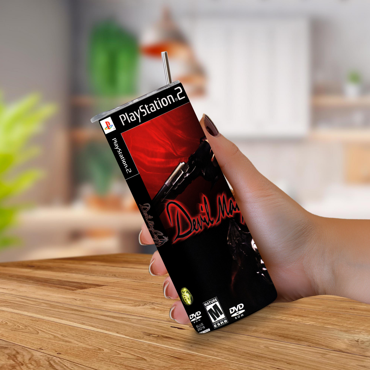 A hand holding a PlayStation 2-themed 20oz stainless steel tumbler with a straw, featuring the 'Devil May Cry' cover art, set against a blurred kitchen background.