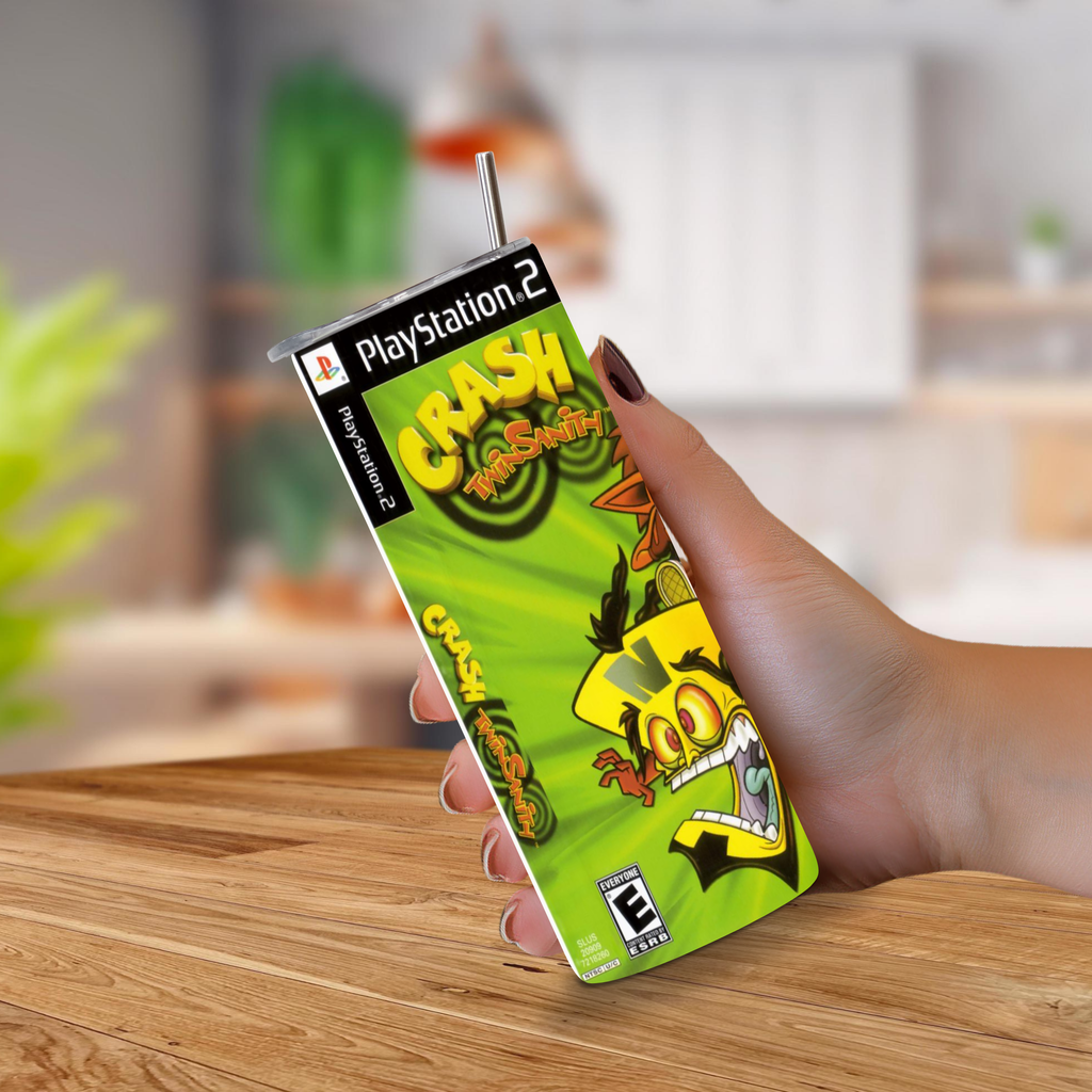 A hand holding a PlayStation 2 Crash Twinsanity game cover designed to look like a 20oz stainless steel tumbler, with a metal straw sticking out of the top, set against a blurred indoor background.