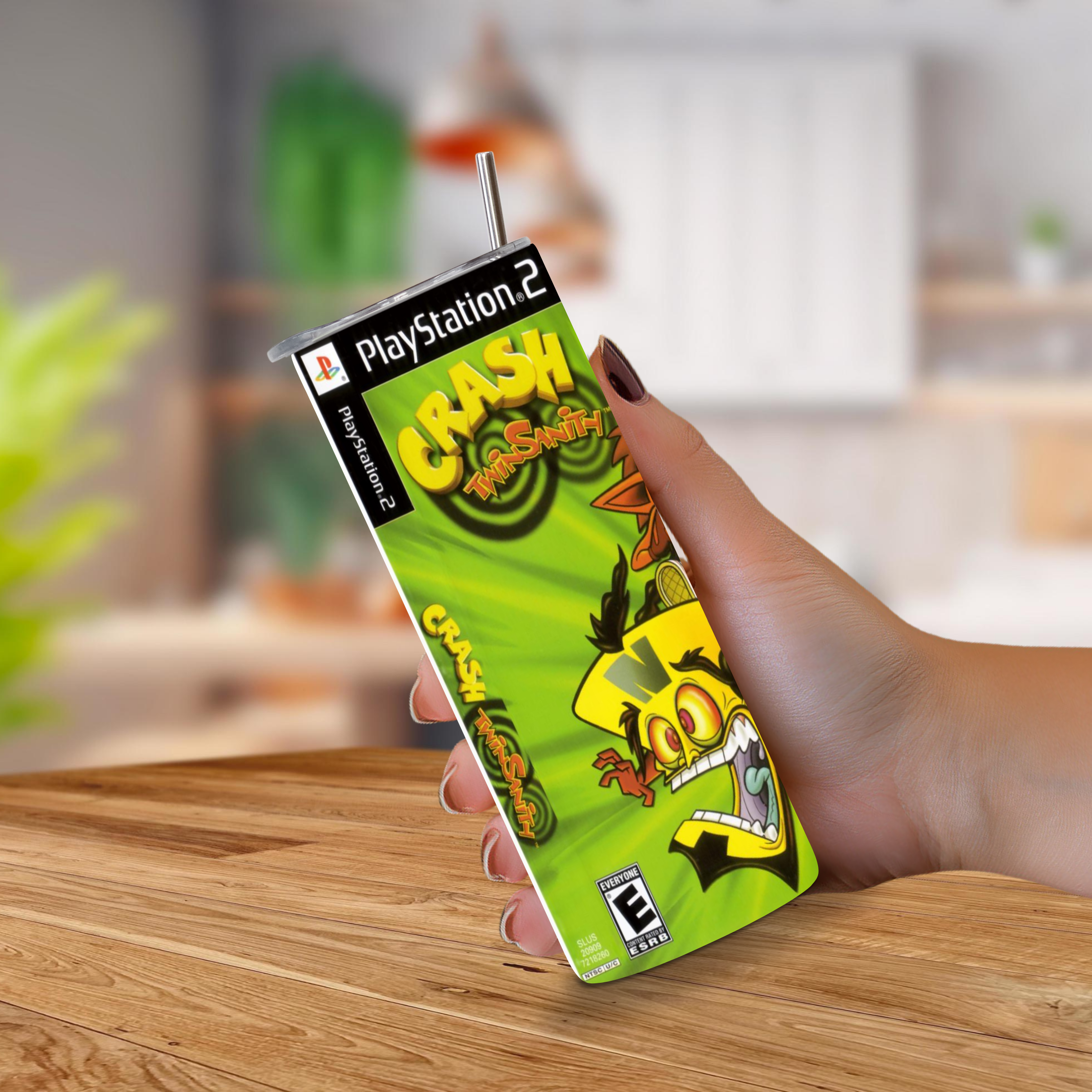 A hand holding a PlayStation 2 Crash Twinsanity game cover designed to look like a 20oz stainless steel tumbler, with a metal straw sticking out of the top, set against a blurred indoor background.