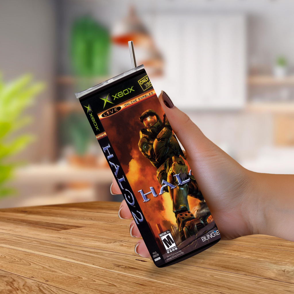 A hand holding a custom-printed tumbler cup designed to look like Xbox Halo 2 game packaging, with a metal straw, set against a blurred kitchen background.