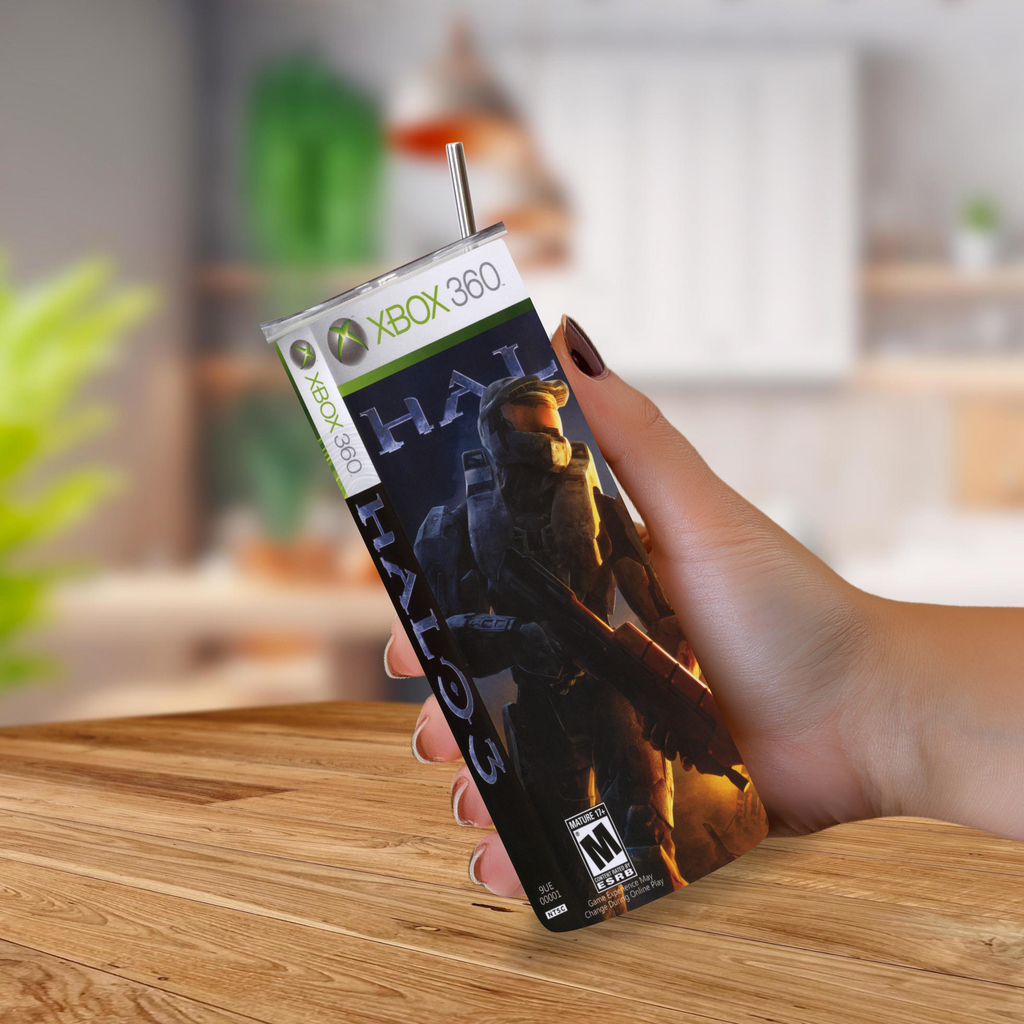 A hand holding a custom-designed tumbler cup that looks like an Xbox 360 Halo 3 Games game case, complete with a metal straw, set against a blurred kitchen background.