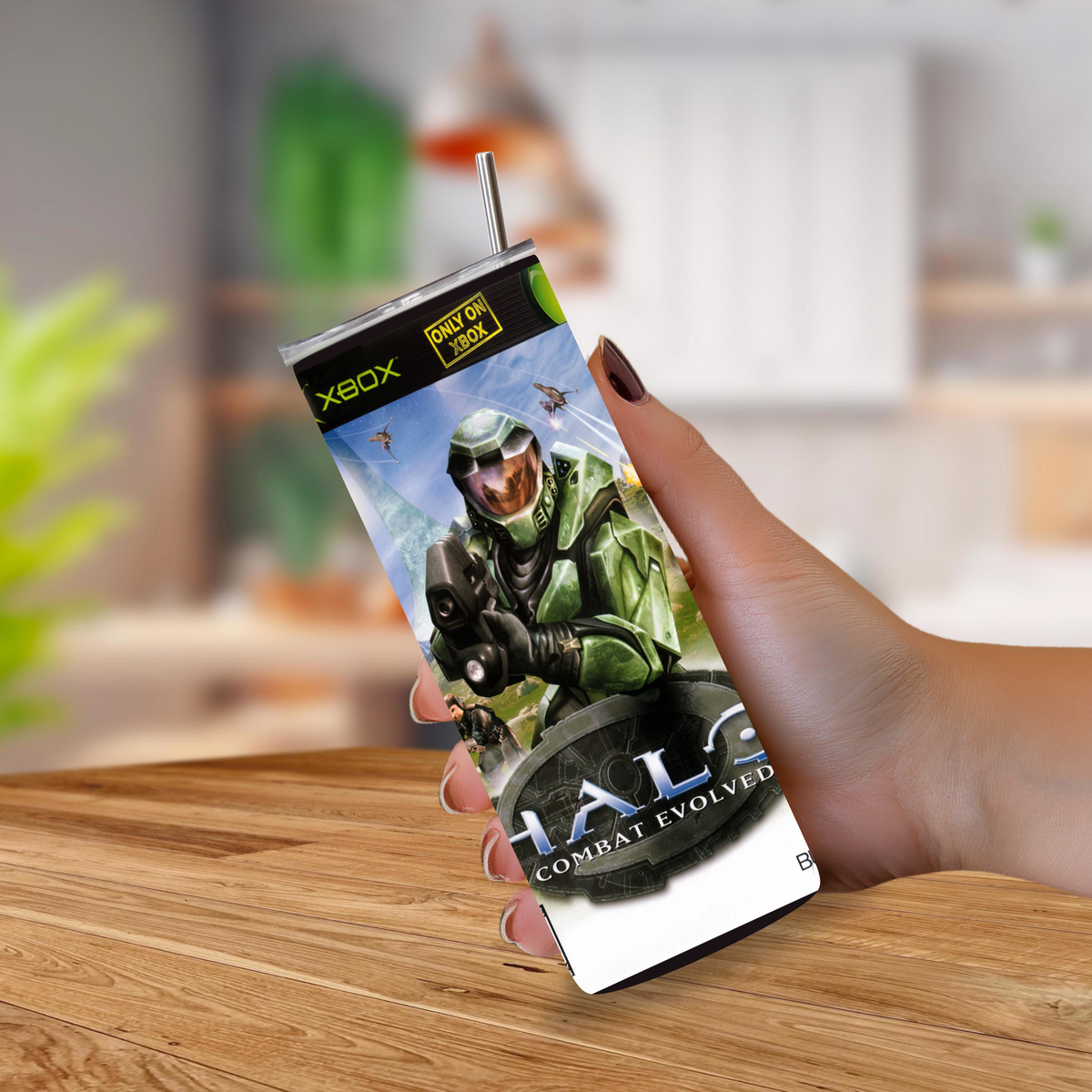 A hand holding a slim rectangular Halo: Combat Evolved themed drink tumbler with a straw, shown against a blurred indoor background with a wooden table in front.