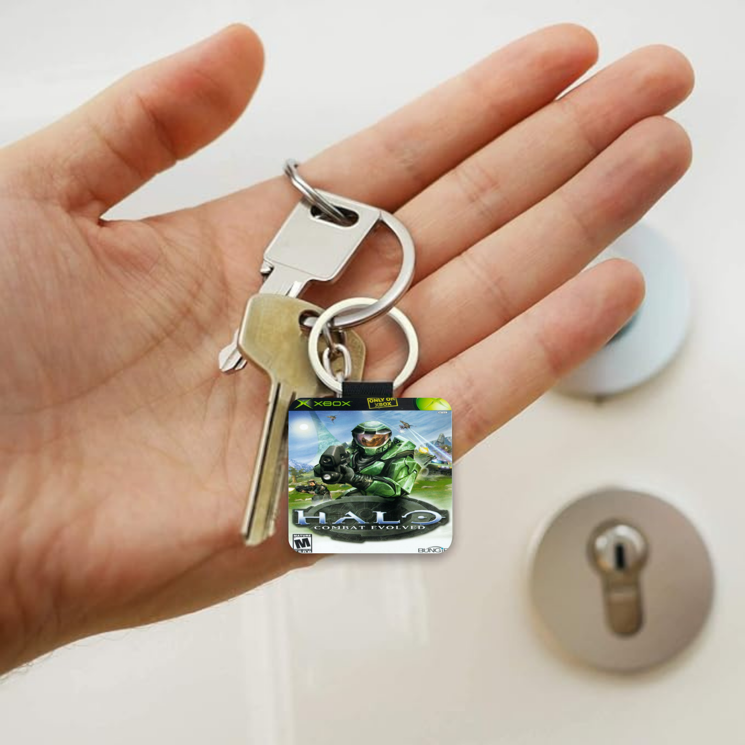 A hand holding a keychain with a square Halo Combat Evolved character charm and two metal keys, shown in front of a lock on a light-colored door.