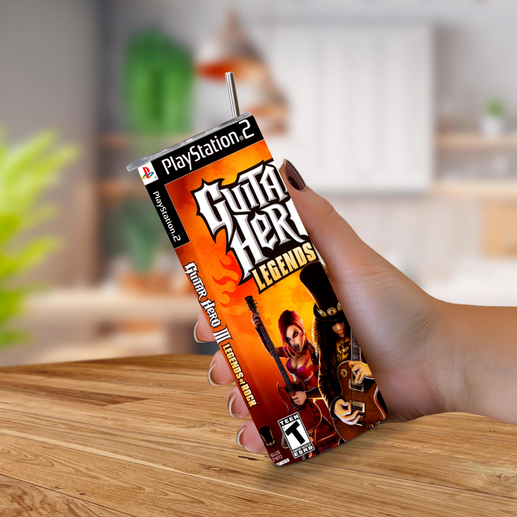 A hand holding a 20oz stainless steel designed to look like a PlayStation 2 game cover, featuring Guitar Hero III: Legends artwork, on a wooden table with a blurred indoor background.