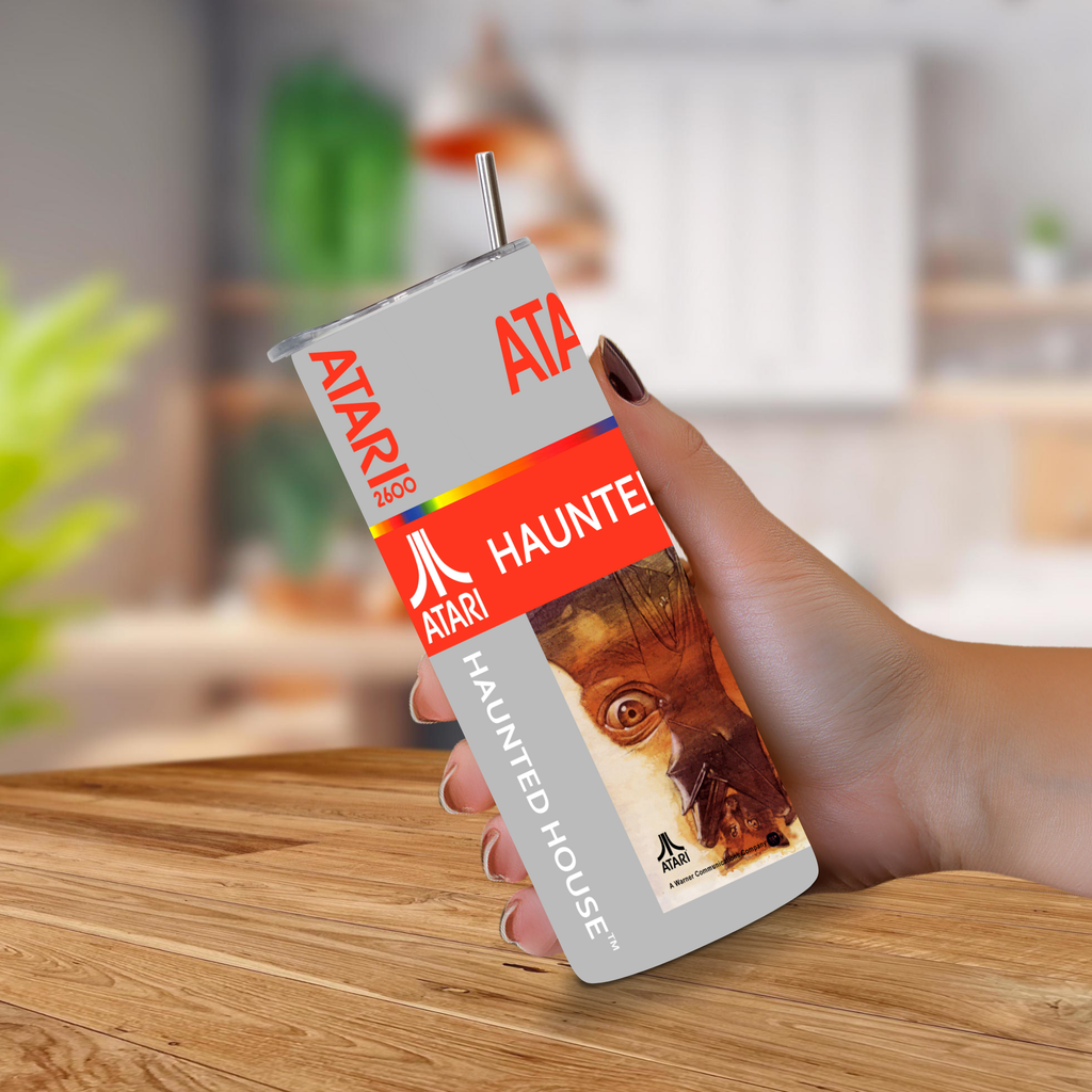 A hand holding a novelty can-shaped tumbler featuring Atari's 'Haunted House' design, set against a blurred kitchen background.