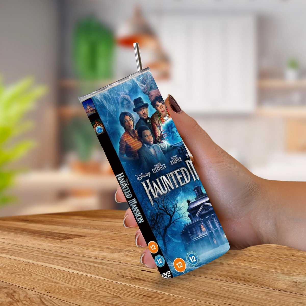 A hand holding a slim 20oz tumbler of Disney's Haunted Mansion, with a metal straw sticking out from the top, set against a blurred indoor background and wooden tabletop.