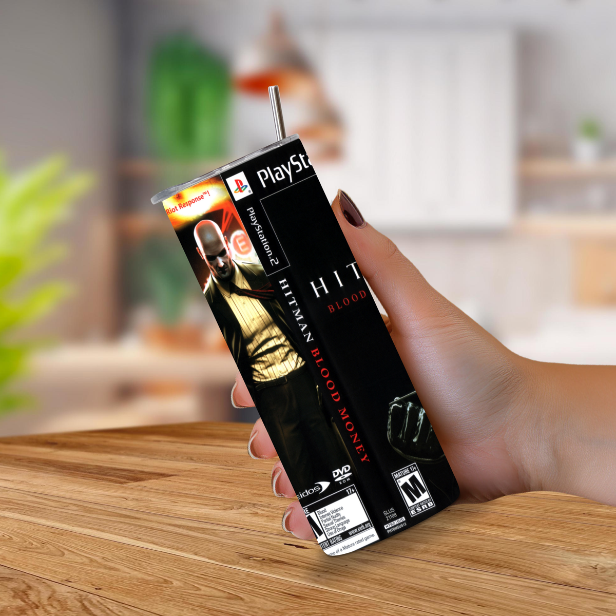 A hand holding a 20oz tumbler styled to look like a PlayStation 2 game case, featuring the Hitman 'Blood Money' cover art, on a wooden table with a blurred kitchen background.