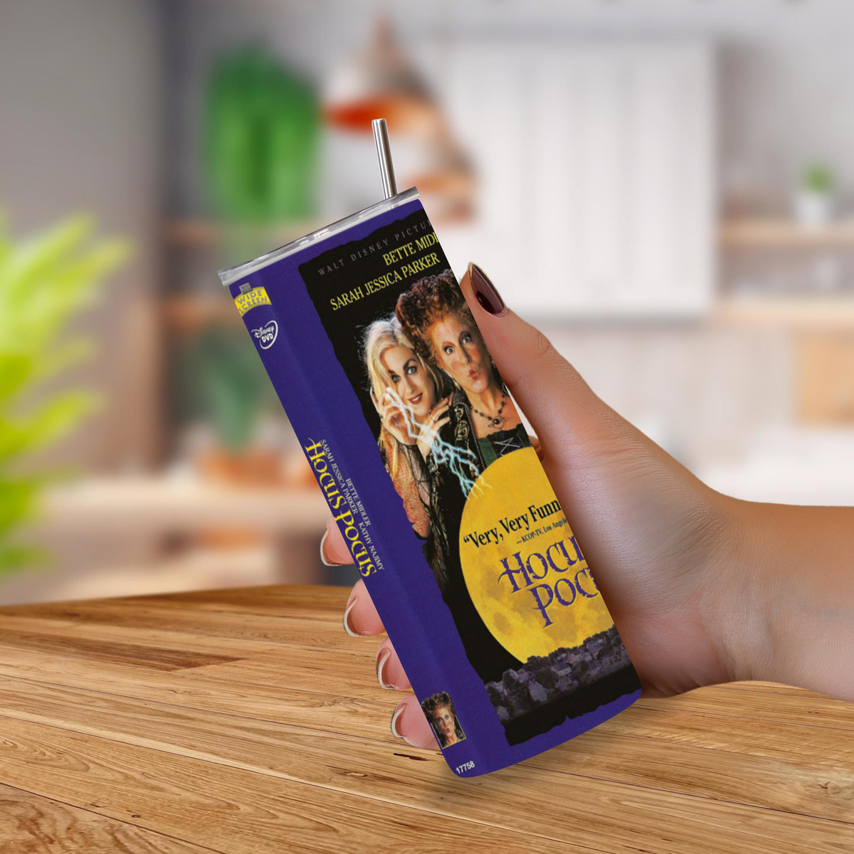 A hand holding a purple and black 'Hocus Pocus' themed 20oz tumbler with a straw, set against a blurred kitchen background
