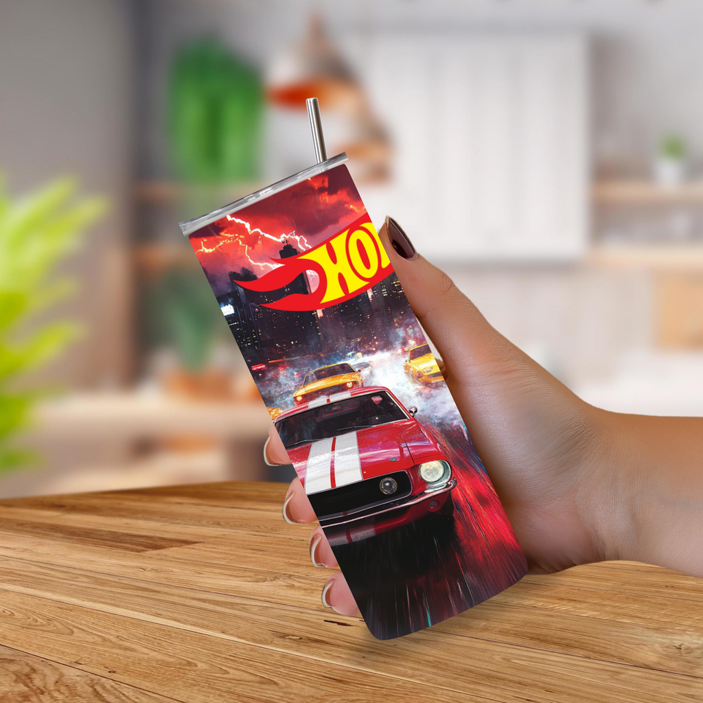 A hand holding a tall insulated tumbler with a printed racing Hot Wheels graphic and a metal straw, photographed in a blurred indoor setting over a wooden table.