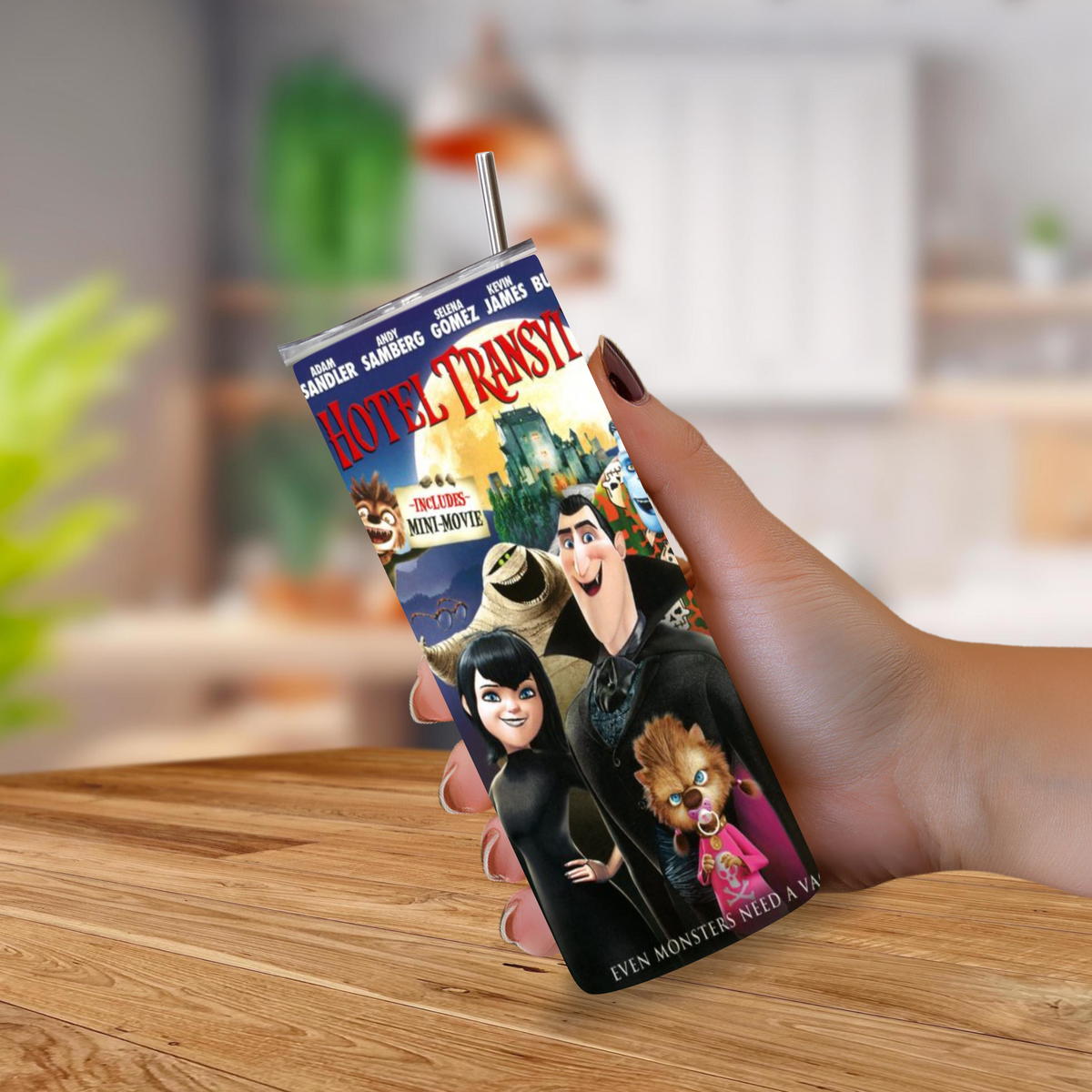 A hand holding a slim, 20oz tumbler with a straw, printed with a colorful Hotel Transylvania movie design, set against a blurred indoor background.