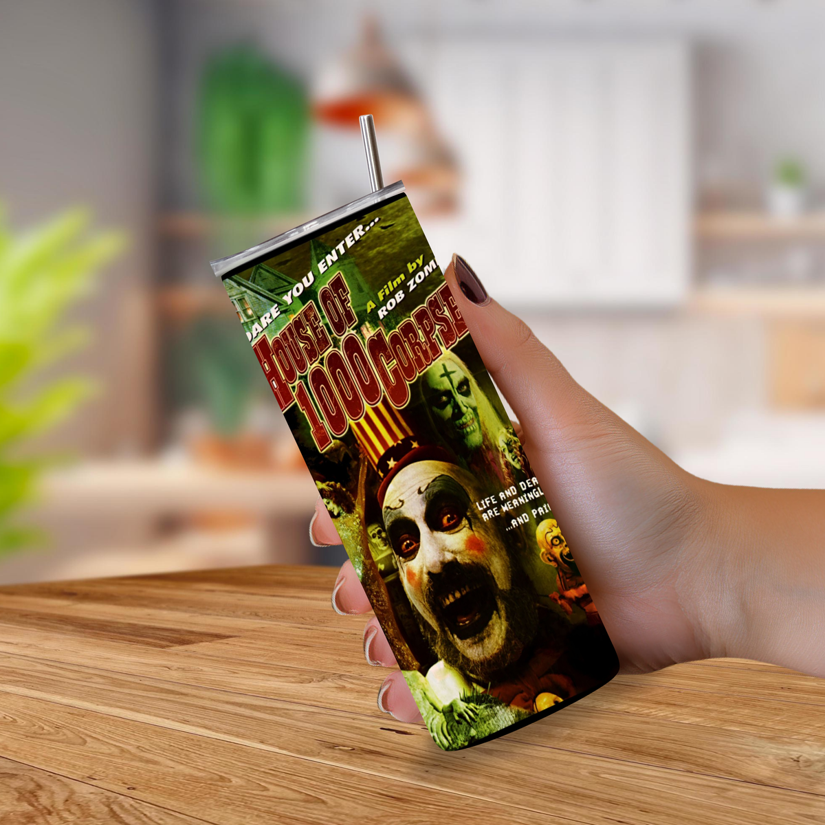 A hand holding a 20oz tumbler featuring movie graphic design from "House of 1000 Corpses" on a blurred background. 