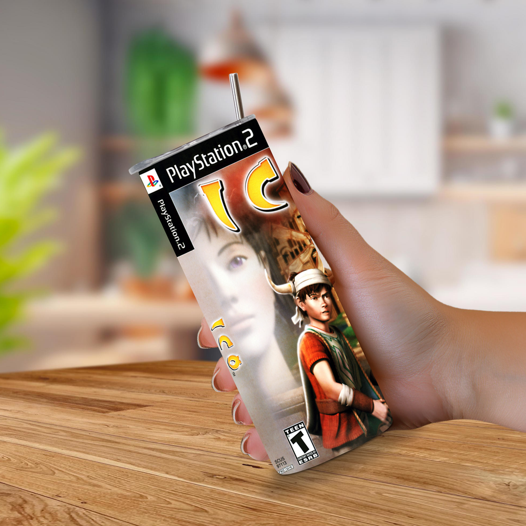 A hand holding a 20oz tumbler with PlayStation 2 ICO video game with a metal straw extending from the top, set against a blurred indoor background and wooden tabletop.