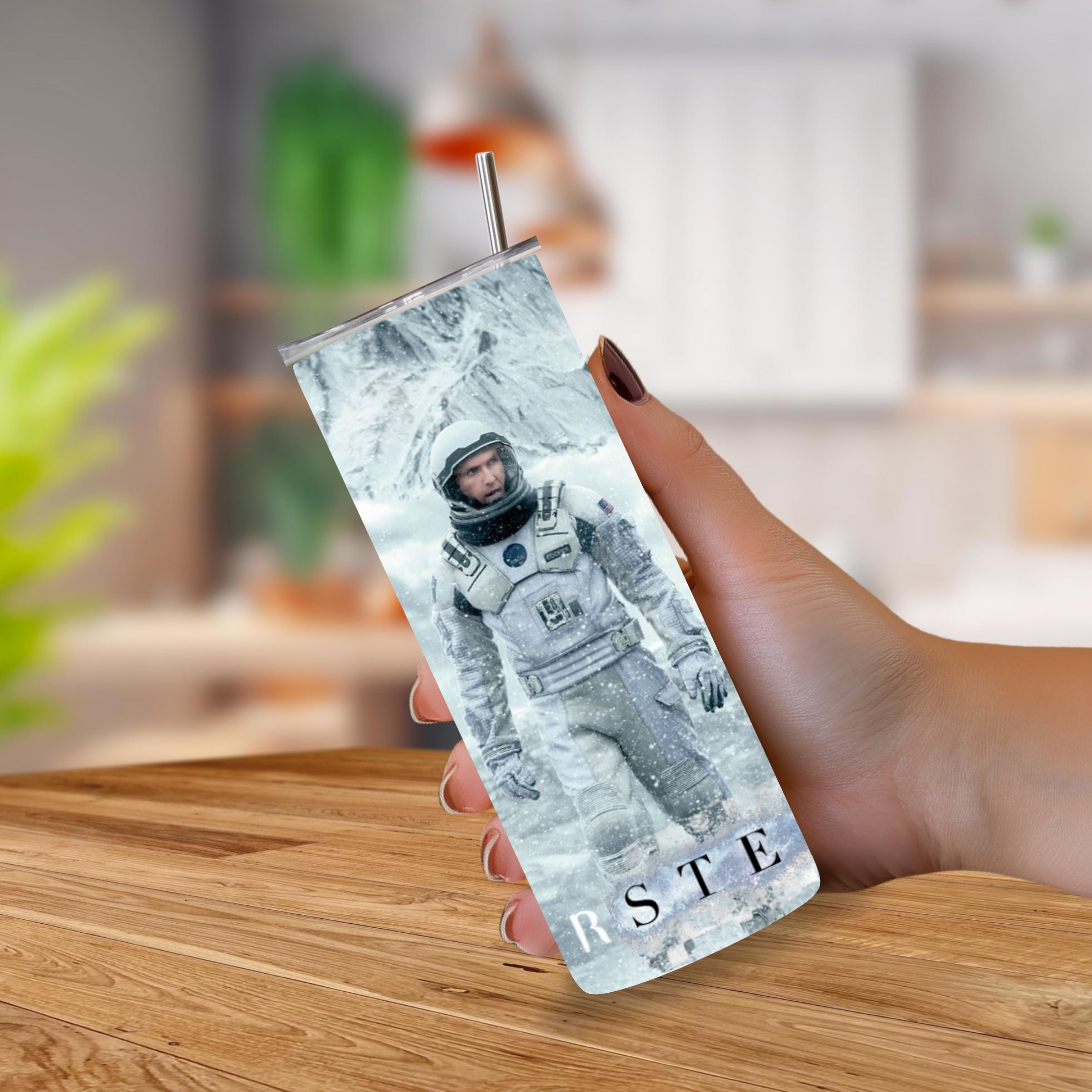 A hand holding a 20oz tumbler with a metal straw, printed with an astronaut graphic and the text 'Interstellar' at the bottom, set against a blurred kitchen background.