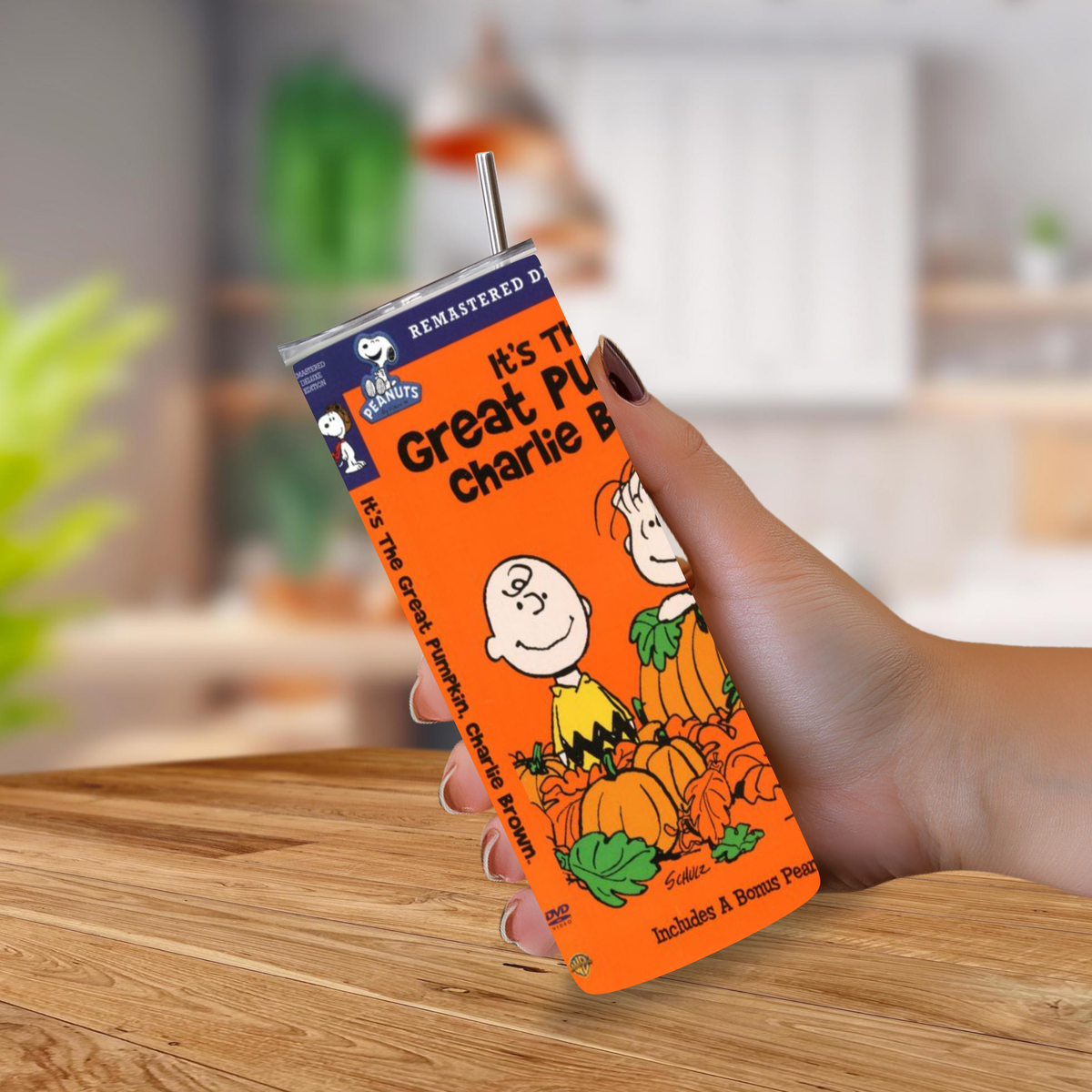 A hand holding a 20oz travel tumbler featuring a remastered Peanuts 'It's the Great Pumpkin Charlie Brown' design, with an orange pumpkin-themed print and a metal straw, set against a blurred kitchen background.