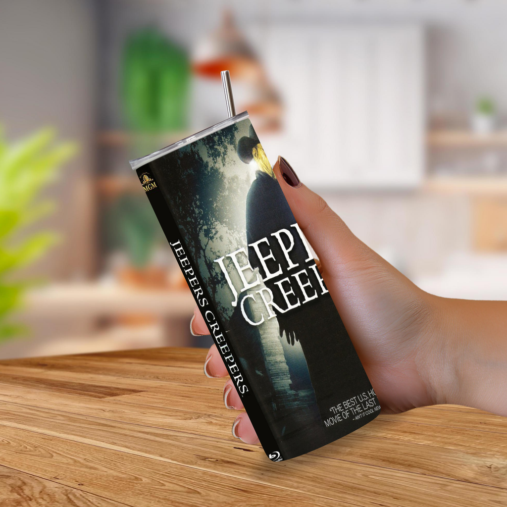 A hand holding a tumbler cup featuring the 'Jeepers Creepers' movie poster artwork, complete with a clear lid and metal straw, set against a blurred kitchen background.