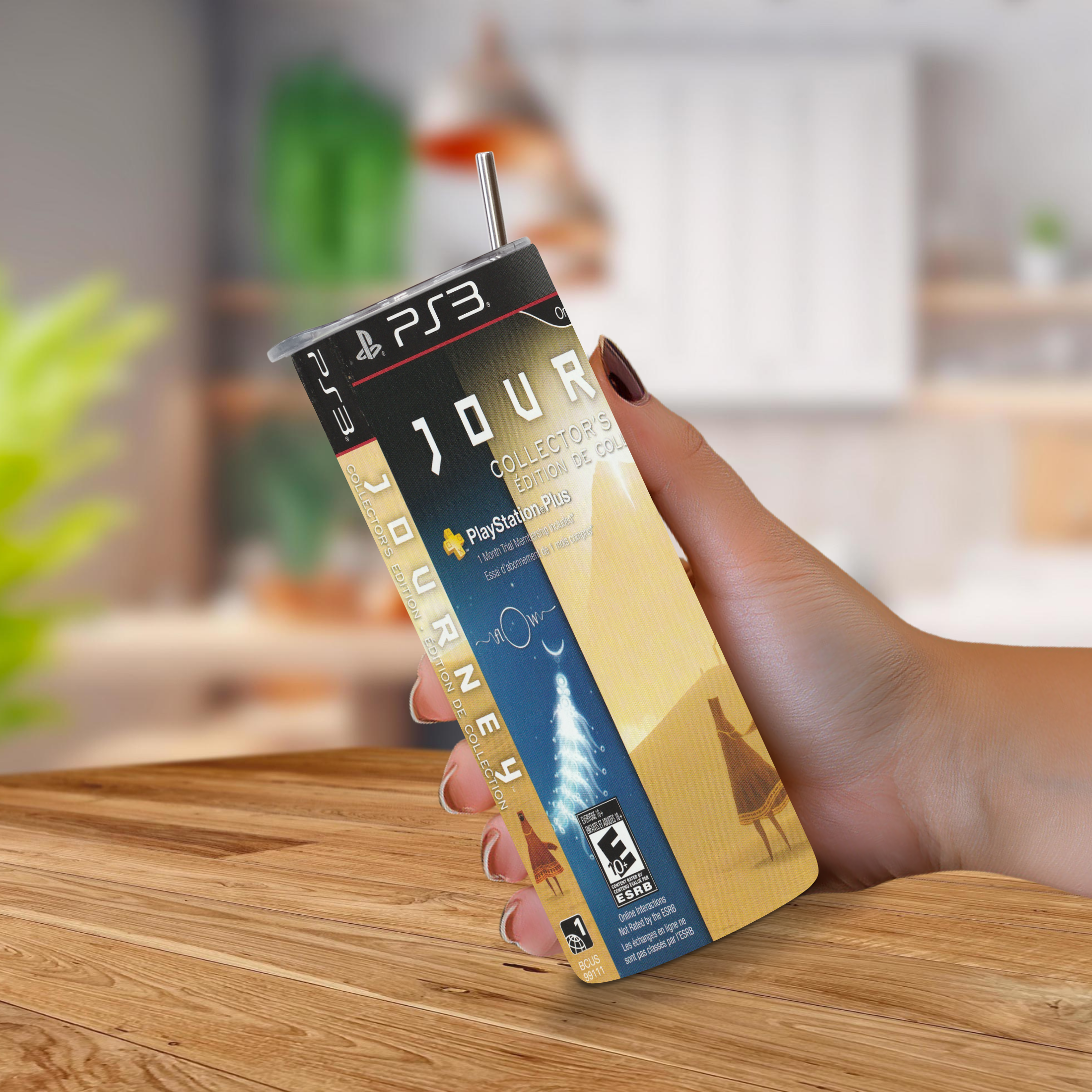 A hand holding a PlayStation 3 themed 20oz stainless steel tumbler featuring the 'Journey' game cover art, set against a blurred indoor background with a wooden table.
