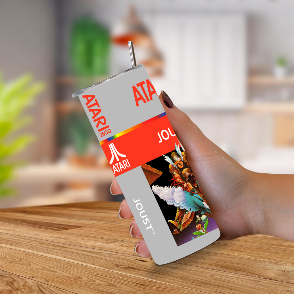 A hand holding a custom-printed Atari-themed tumbler cup with a straw, featuring retro red and white branding and a colorful graphic of Joust, set against a blurred kitchen background.