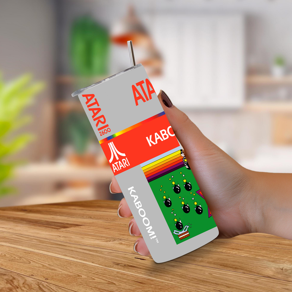 A hand holding a retro-themed 20oz stainless steel tumbler featuring Atari branding and the word KABOOM!, set against a blurred kitchen background.