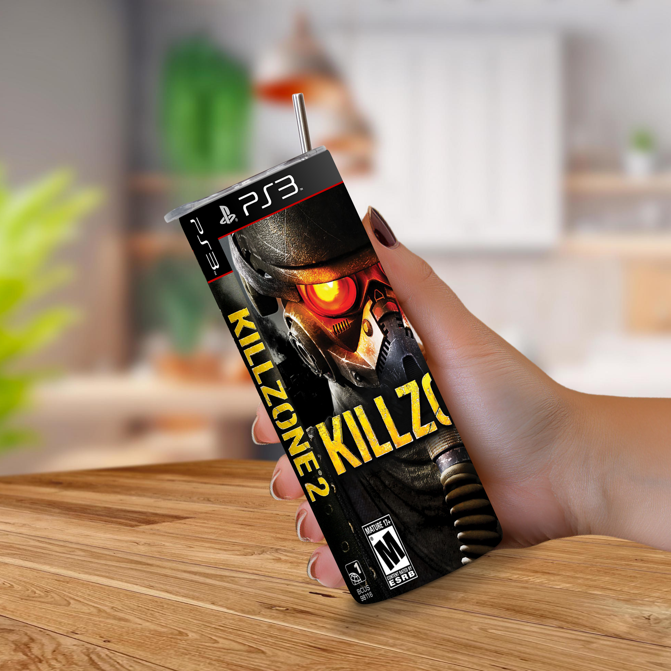 A hand holding a PS3-themed 20oz stainless steel tumbler mockup featuring Killzone 2 artwork, with a straw on top, set against a blurred indoor background and wooden tabletop.