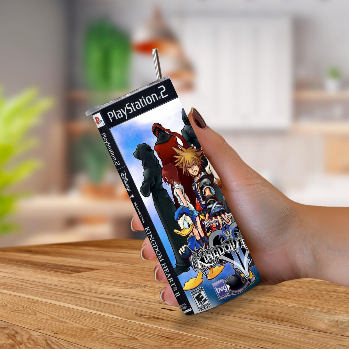 A hand holding a slim PlayStation 2 game 20oz tumbler designed to look like the Kingdom Hearts set against a blurred indoor background.