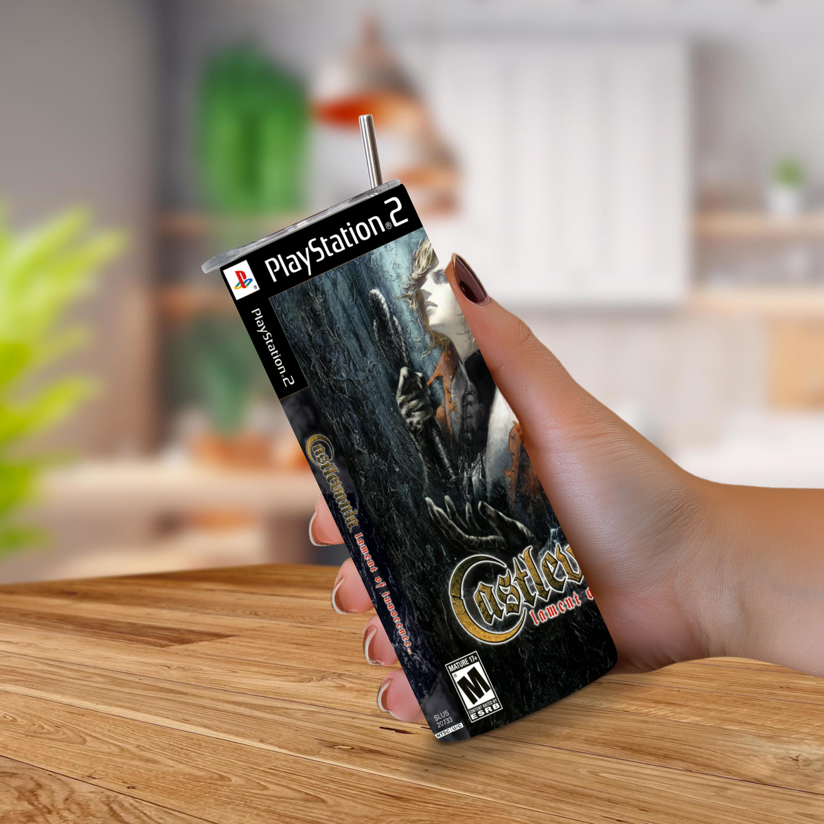 A hand holding a 20oz stainless steel tumbler PlayStation 2 game cover for Castlevania: Lament of Innocence on a wooden tabletop with a blurred kitchen background.