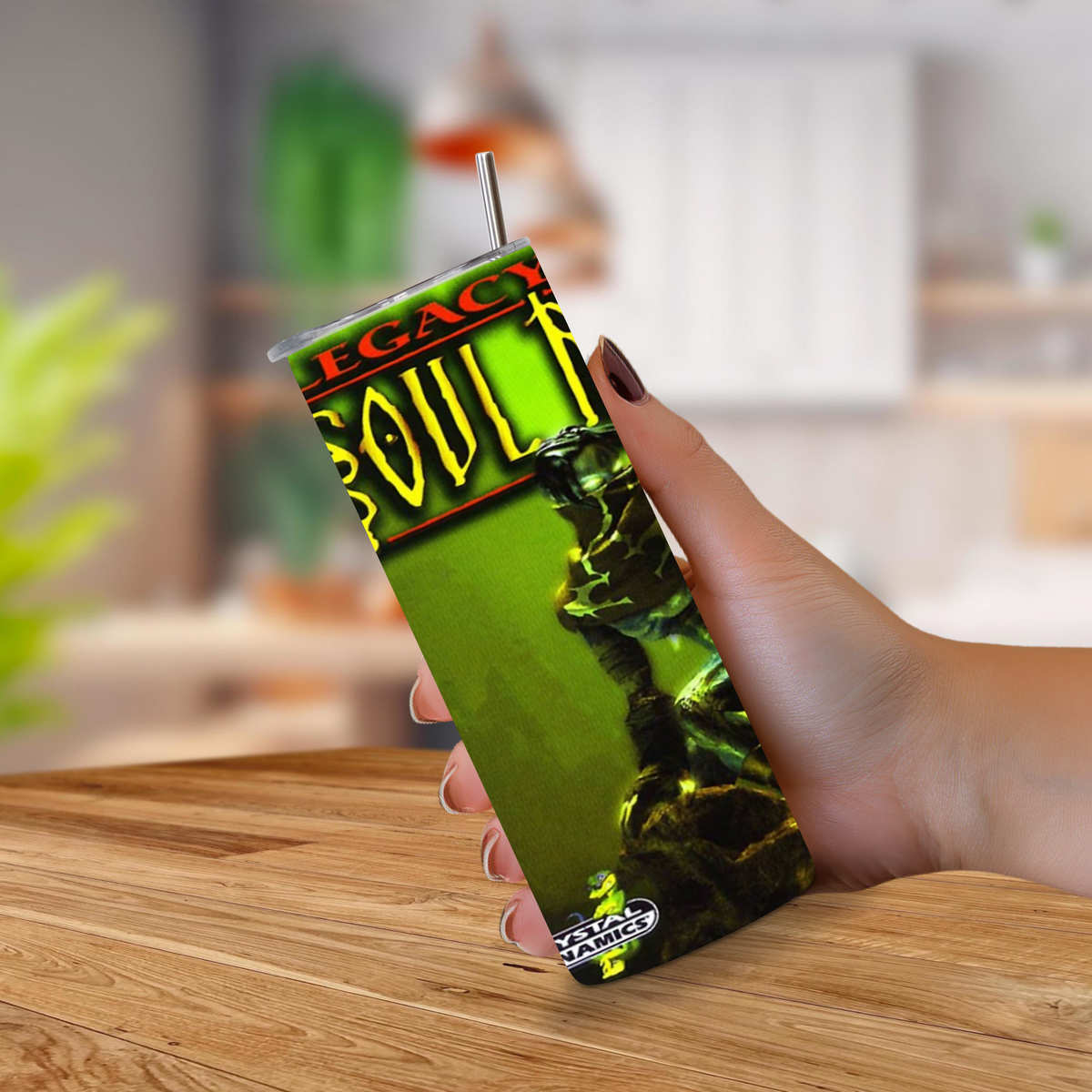 A hand holding a 20oz tumbler with Legacy of Kain PlayStation graphics with a metal straw, set against a blurred kitchen background.