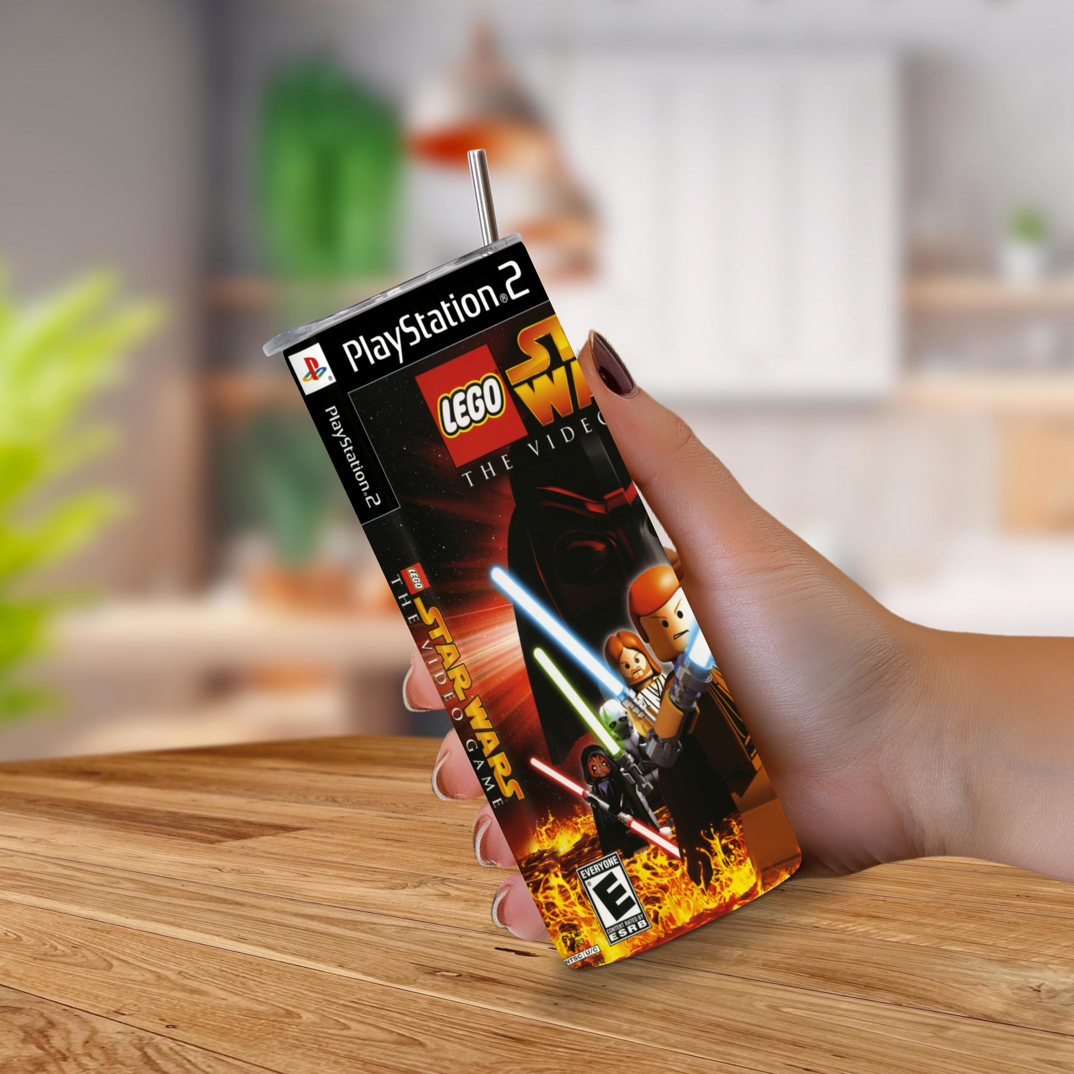 A hand holding a 20oz tumbler featuring LEGO Star Wars: The Video Game artwork, on a wooden table with a blurred indoor background.