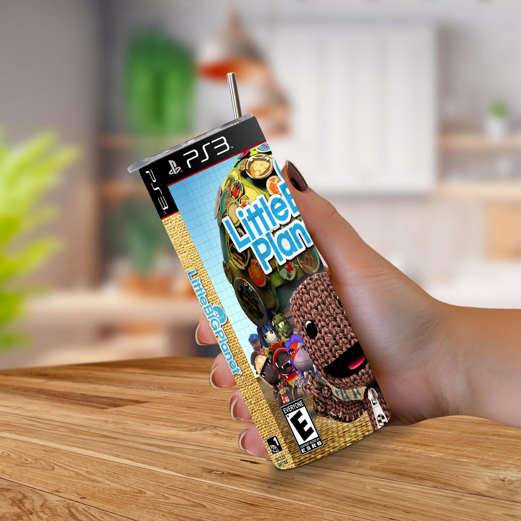 A hand holding a PS3 game case designed to look a 20oz stainless steel tumbler, featuring the 'Little Big Planets' logo and Sackboy artwork, with a metal straw sticking out of the top, set against a blurred kitchen background.