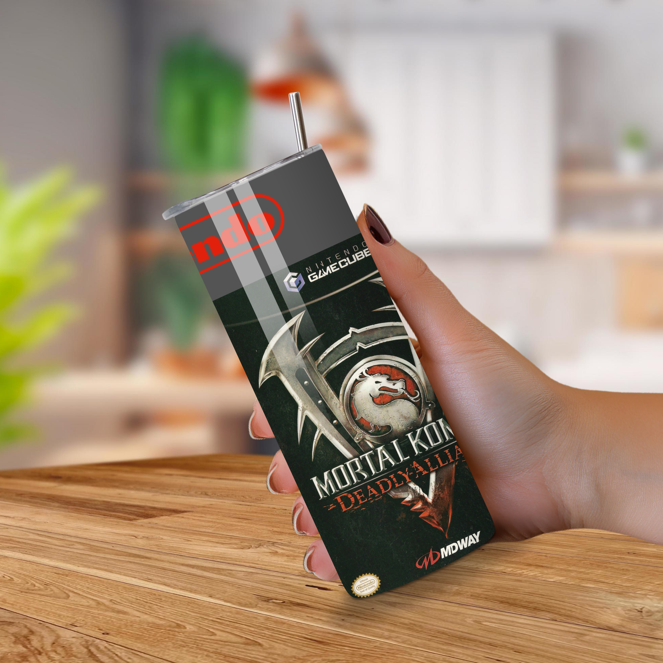 Hand holding a 'Mortal Kombat' GameCube 20oz tumbler on a wooden surface with a blurred background