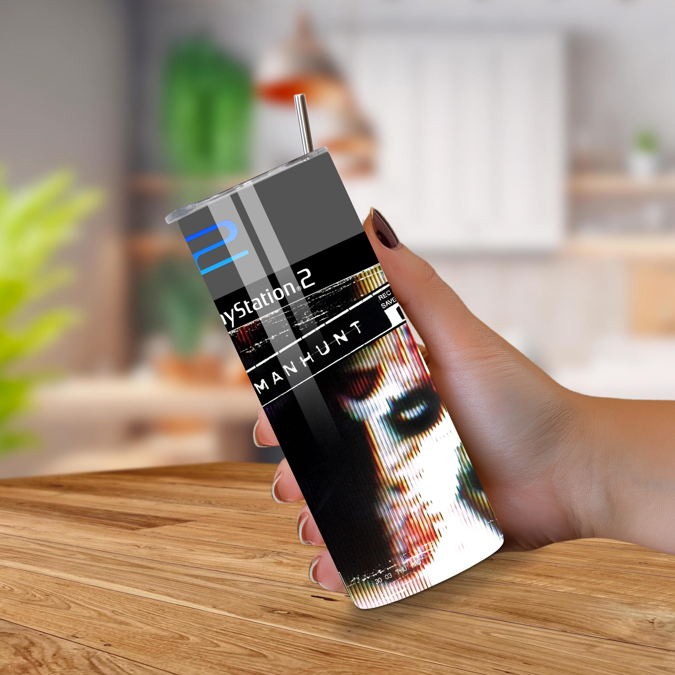 Hand holding a 20oz tumbler with 'PlayStation 2 Manhunt' design on a wooden surface