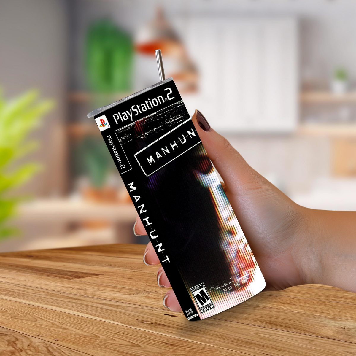 A hand holding a 20oz tumbler featuring the Manhunt PS2 cover art, with a metal straw inserted. The background shows a blurred kitchen setting with a wooden table.