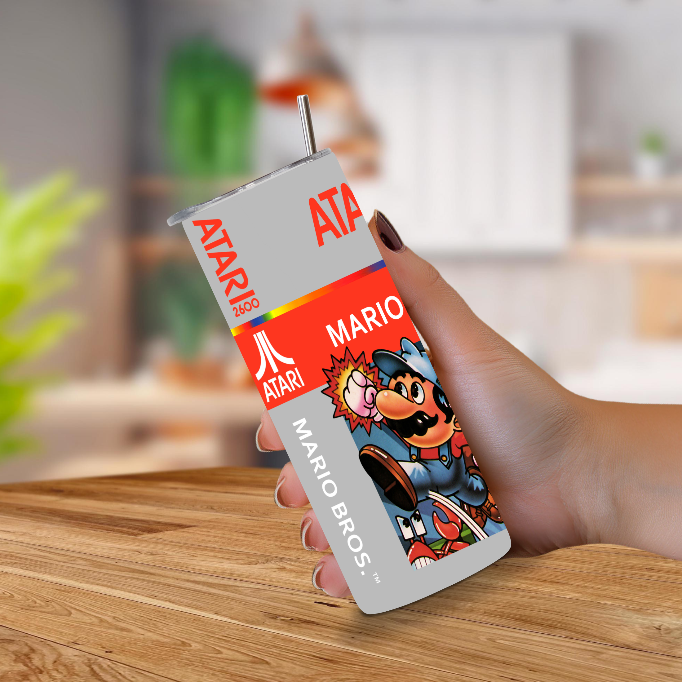 A hand holding a novelty 20oz stainless steel tumbler featuring retro Atari Mario Bros. graphics, set against a blurred kitchen background.
