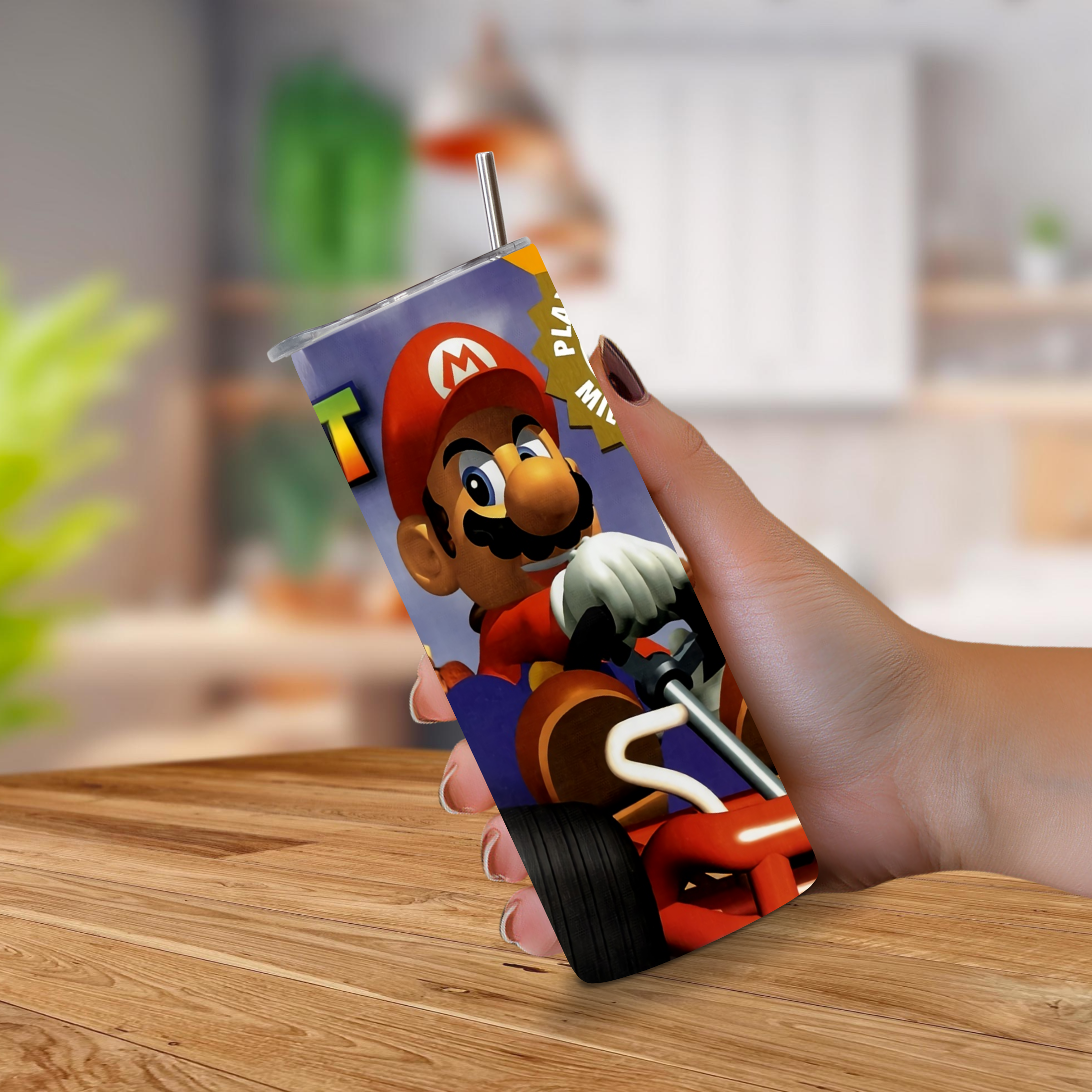 A hand holding a 20oz stainless steel tumbler featuring Mario Kart 64 graphic, set against a blurred indoor background with a wooden table.
