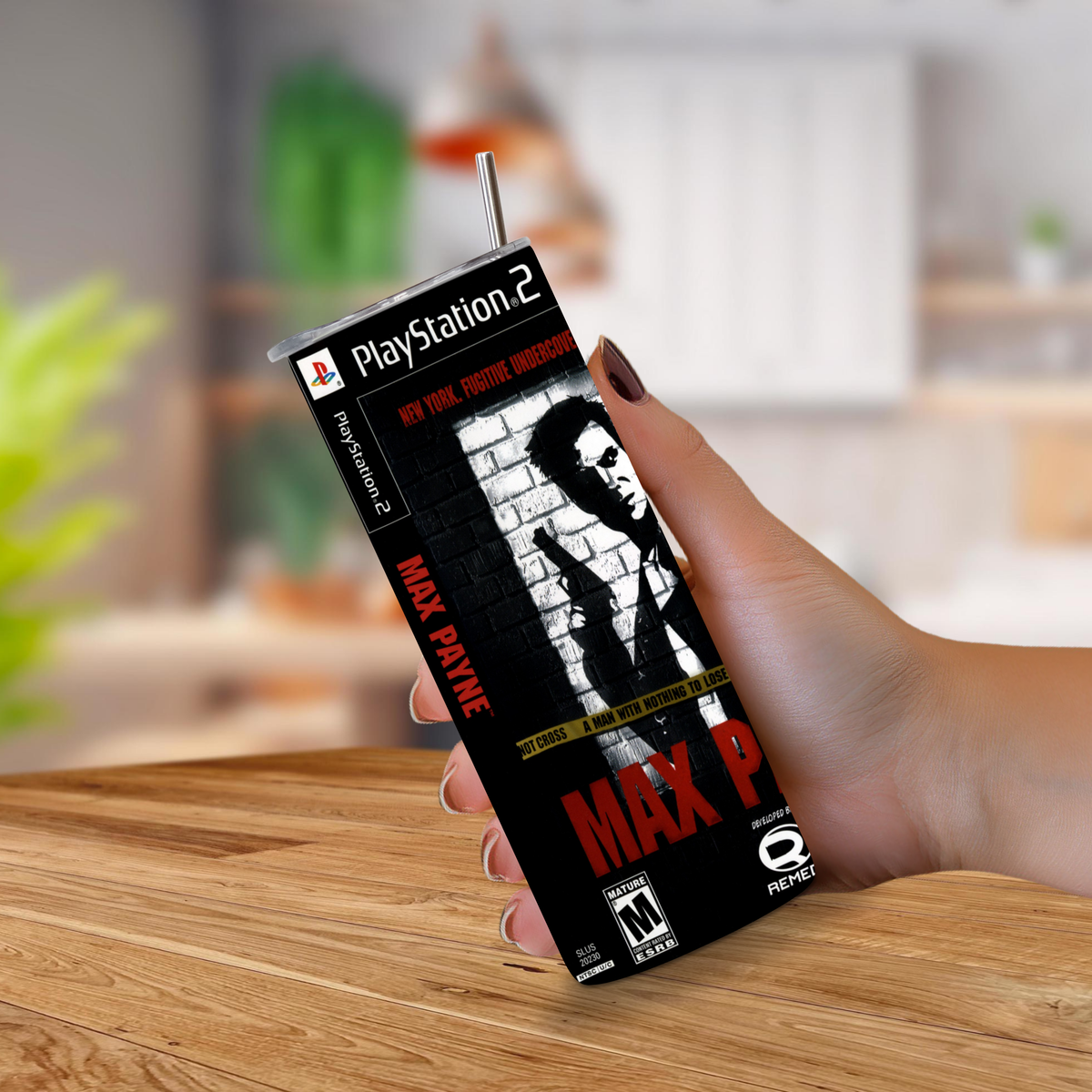 A hand holding a PlayStation 2 Max Payne 20oz tumbler with a straw, set against a blurred indoor background with a wooden table.
