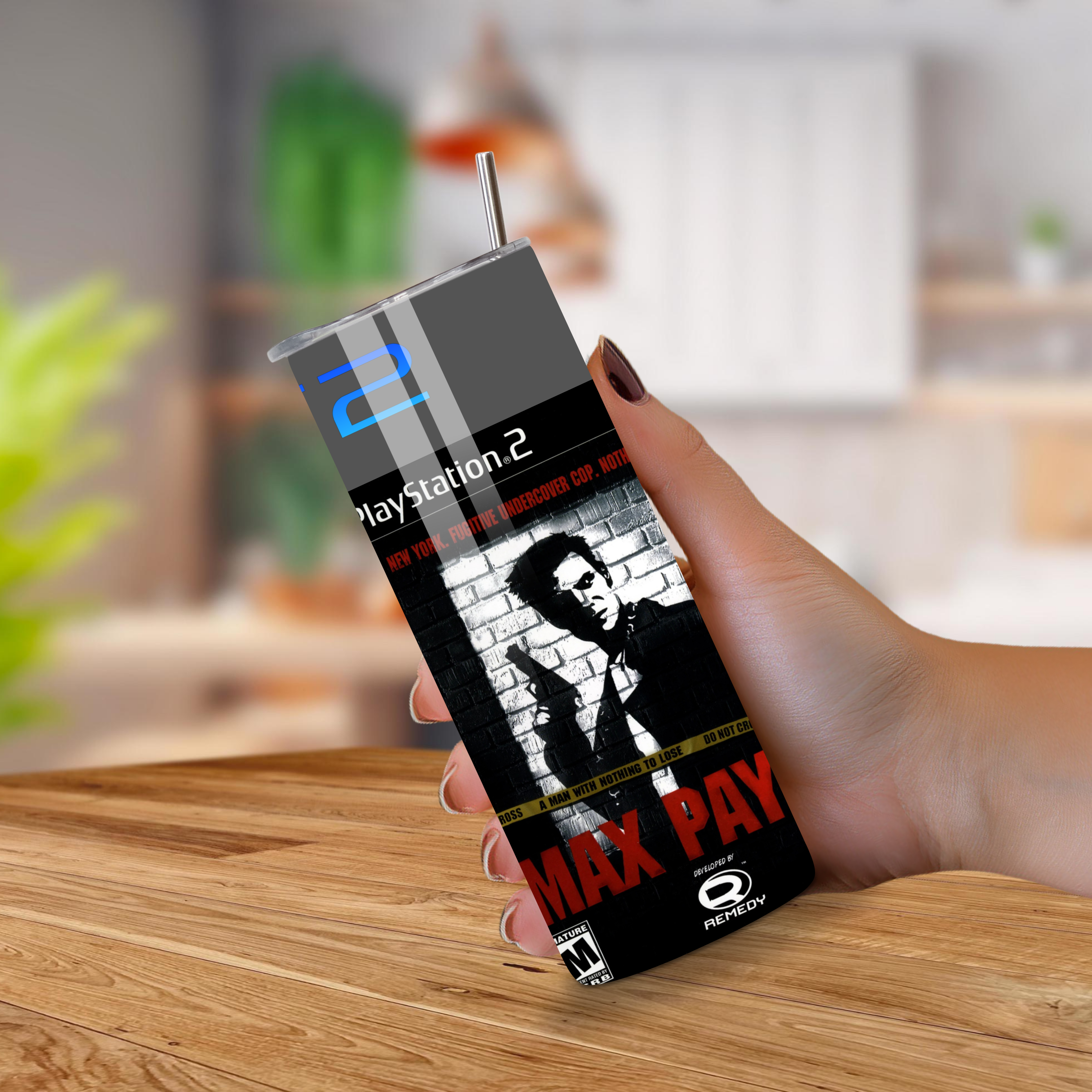 PlayStation 2 tumbler with 'Max Payne' design held by a hand on a wooden surface.