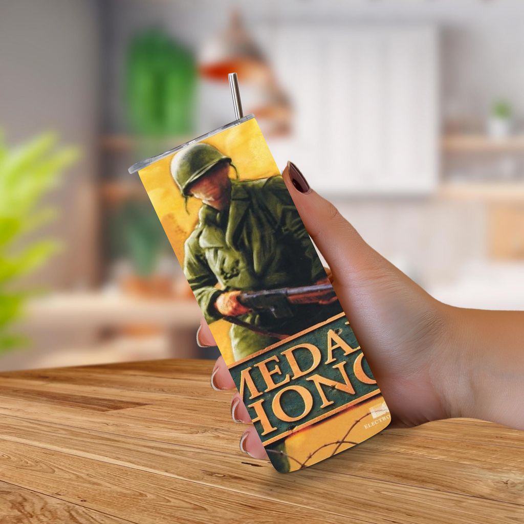A hand holding a 20oz Stainless Steel Tumbler featuring a vintage-style illustration of a soldier in green uniform against a yellow background, with large text reading 'Medal of Honor' near the bottom. The cup has a clear lid and a metal straw, and the background shows a softly blurred indoor setting with a wooden table.