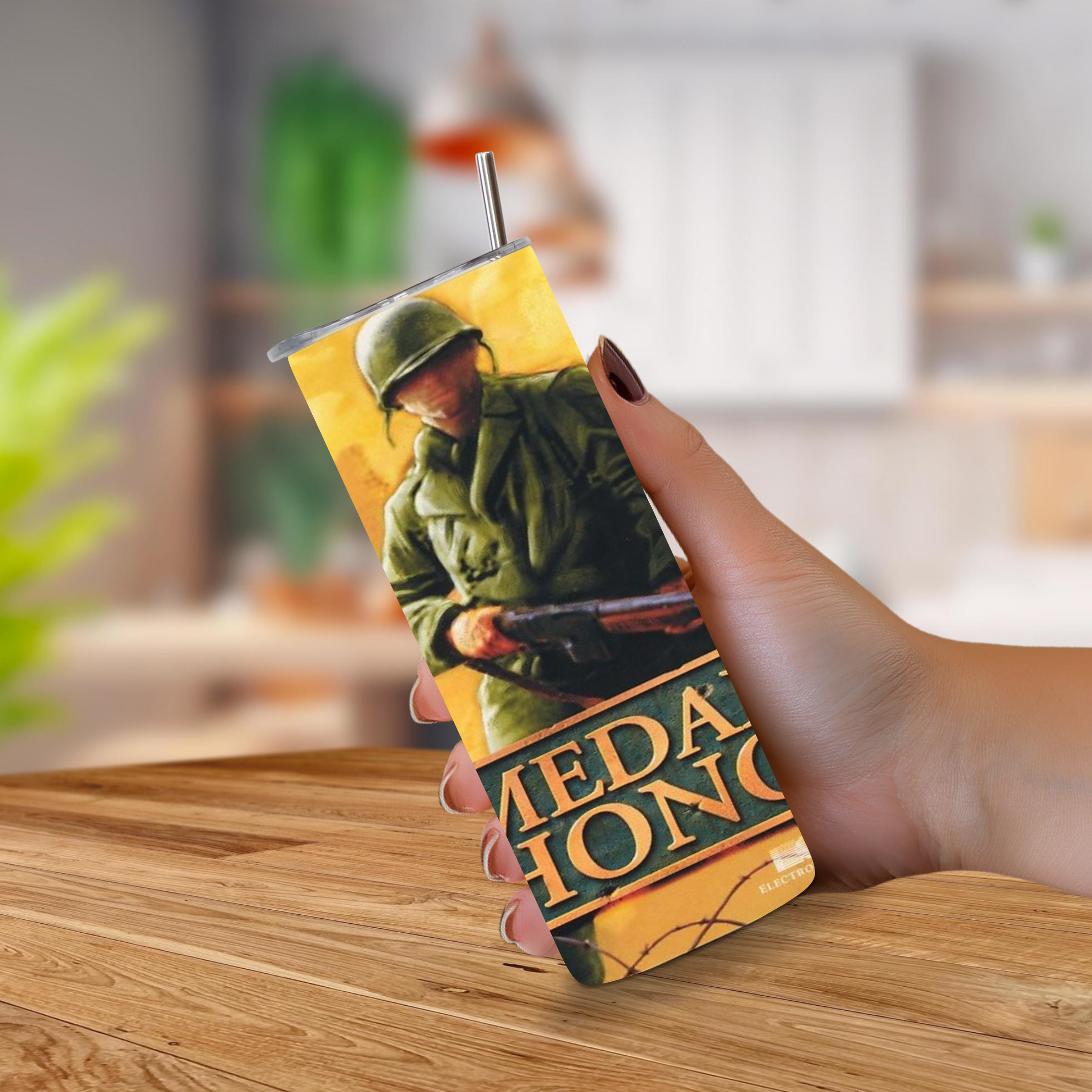 A hand holding a 20oz Stainless Steel Tumbler featuring a vintage-style illustration of a soldier in green uniform against a yellow background, with large text reading 'Medal of Honor' near the bottom. The cup has a clear lid and a metal straw, and the background shows a softly blurred indoor setting with a wooden table.