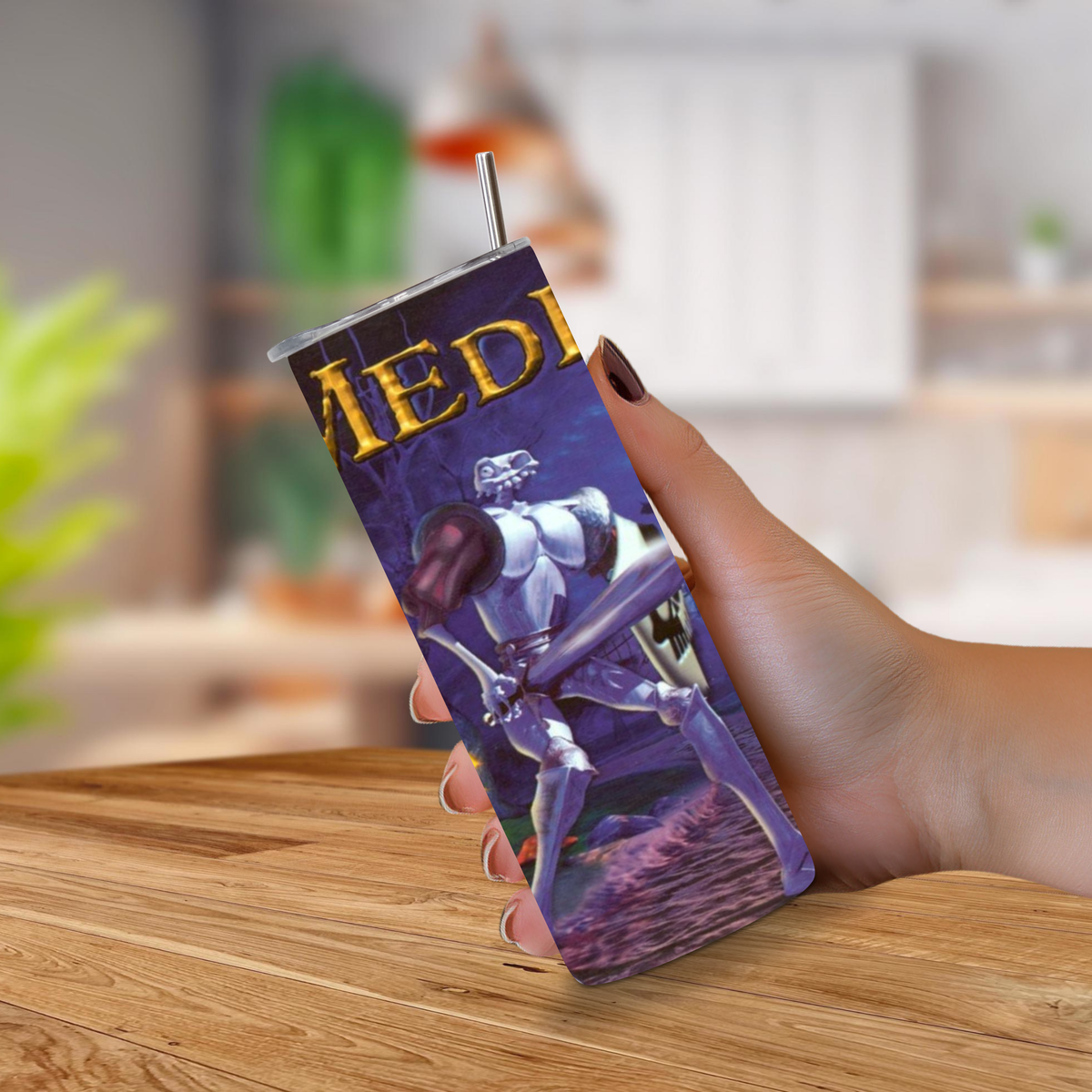 A hand holding a 20oz tumbler with a printed PS1 fantasy illustration featuring MediEvil graphics in gold at the top, set against a blurred indoor background with a wooden table in the foreground.