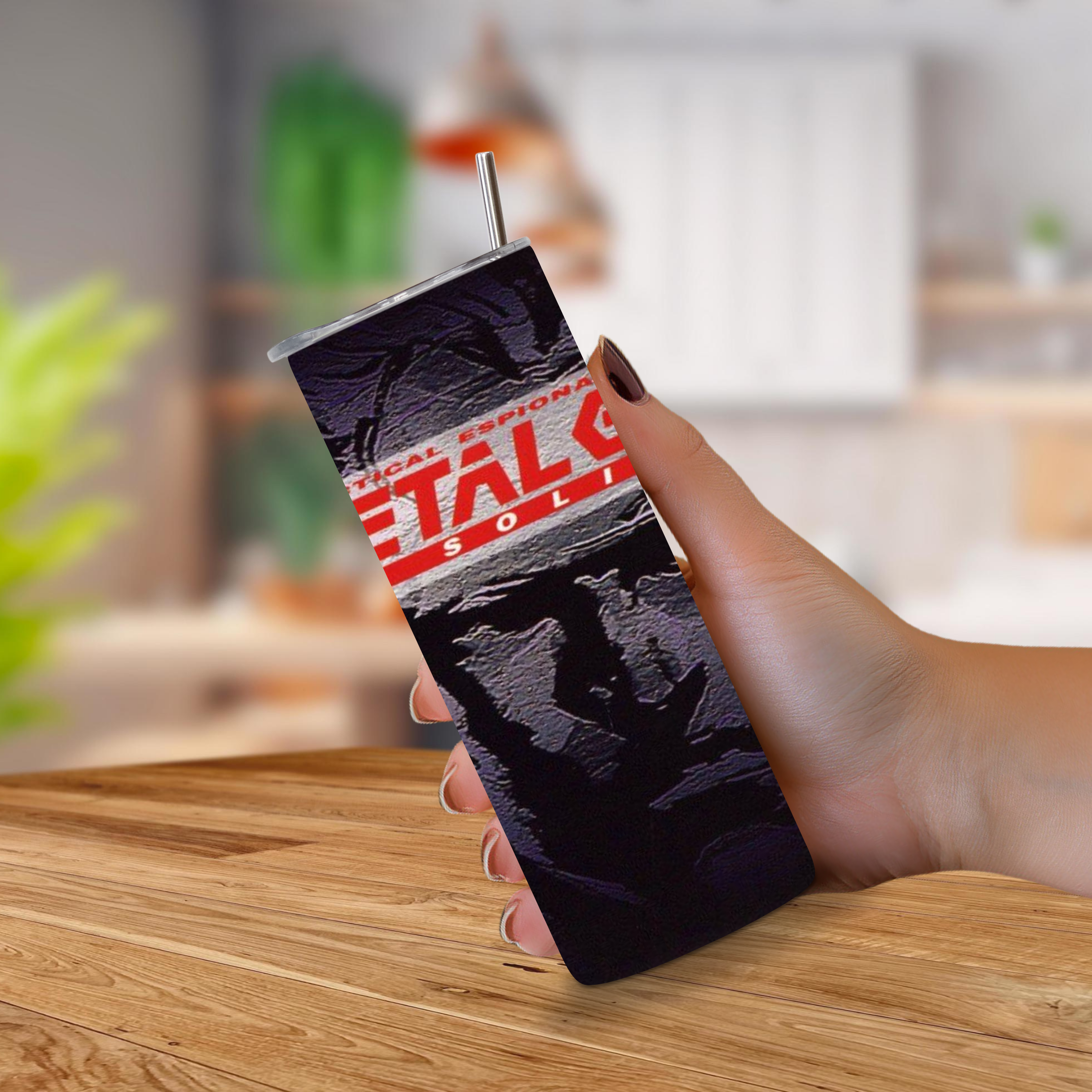 A hand holding a 20oz tumbler with a straw, featuring a dark textured design with red and white 'METAL GEAR SOLID' text, set against a blurred kitchen background.