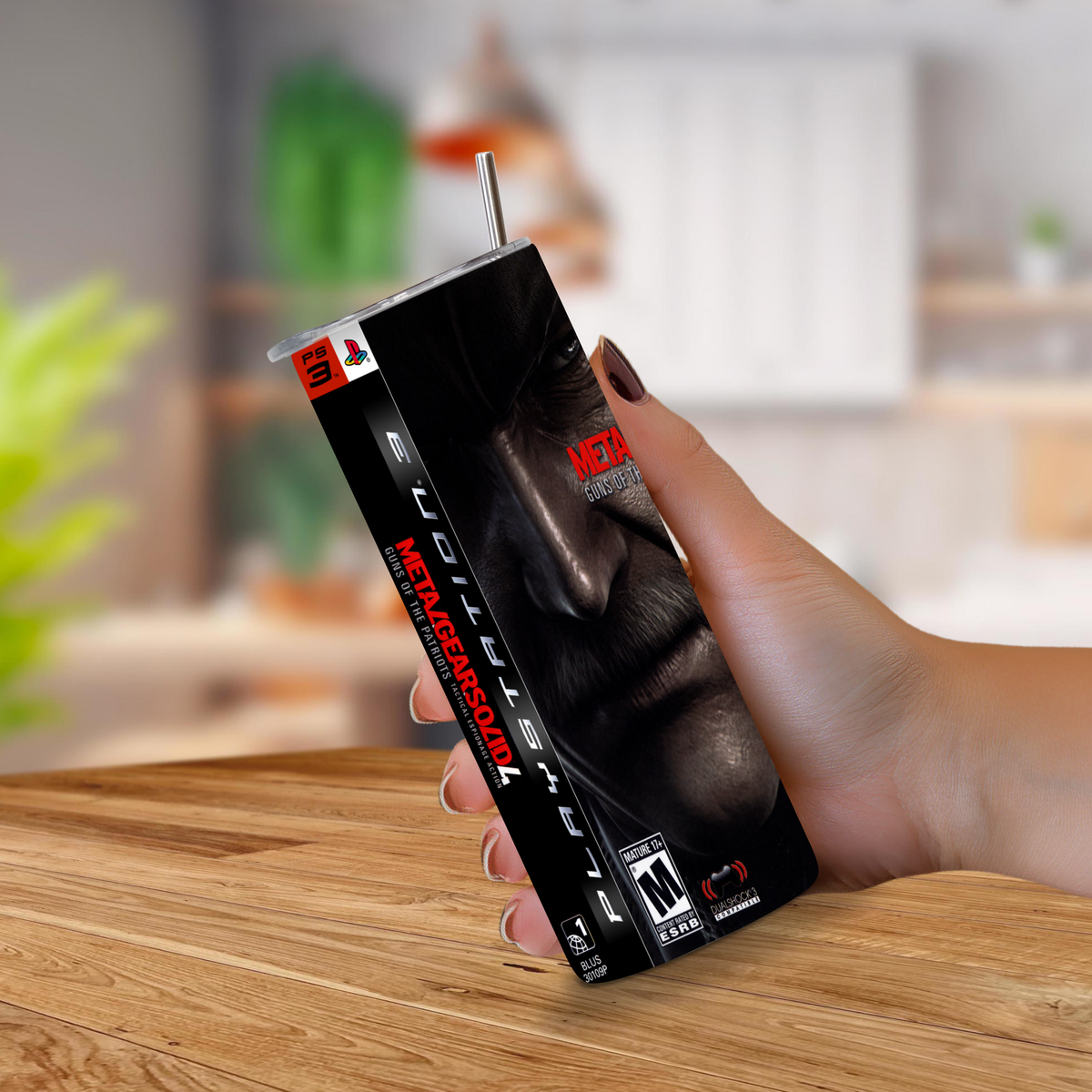 A hand holding 20oz stainless steel metal gear solid 4 guns of the patriots tumbler with a ps3 theme, background of a blurred kitchen.