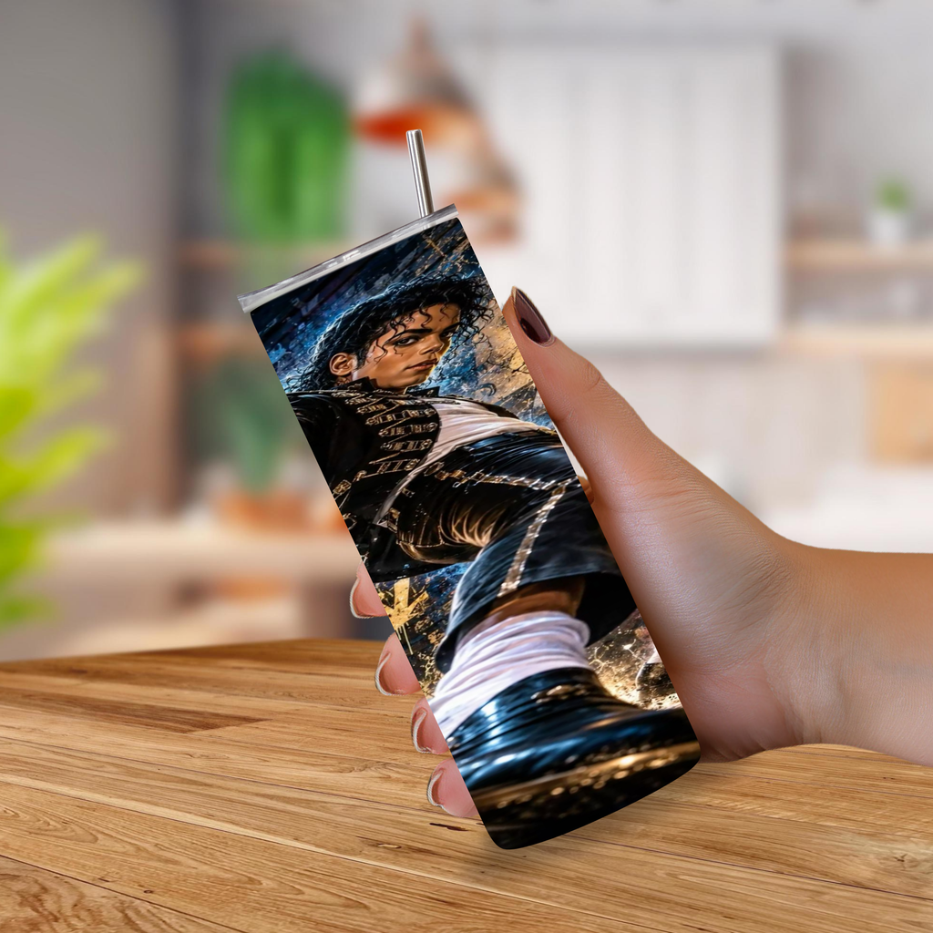 A hand holding a slim stainless steel tumbler with a metal straw, featuring a printed graphic of Michael Jackson in a black jacket and white socks. The tumbler is shown against a blurred indoor background with a wooden table in the foreground.