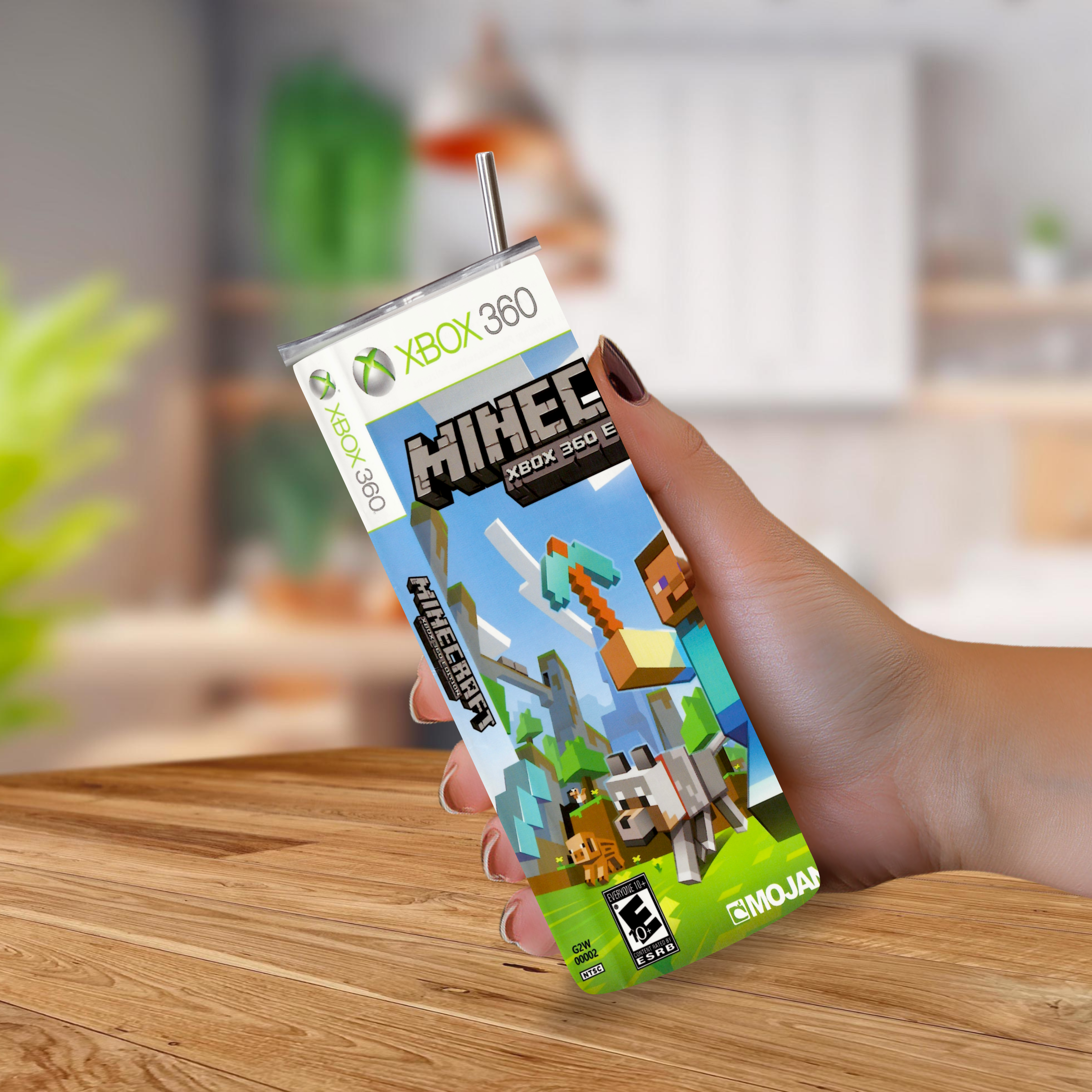 A hand holding a 20oz tumbler styled to look like Minecraft Xbox 360 game packaging, with a straw and a blurred indoor background.