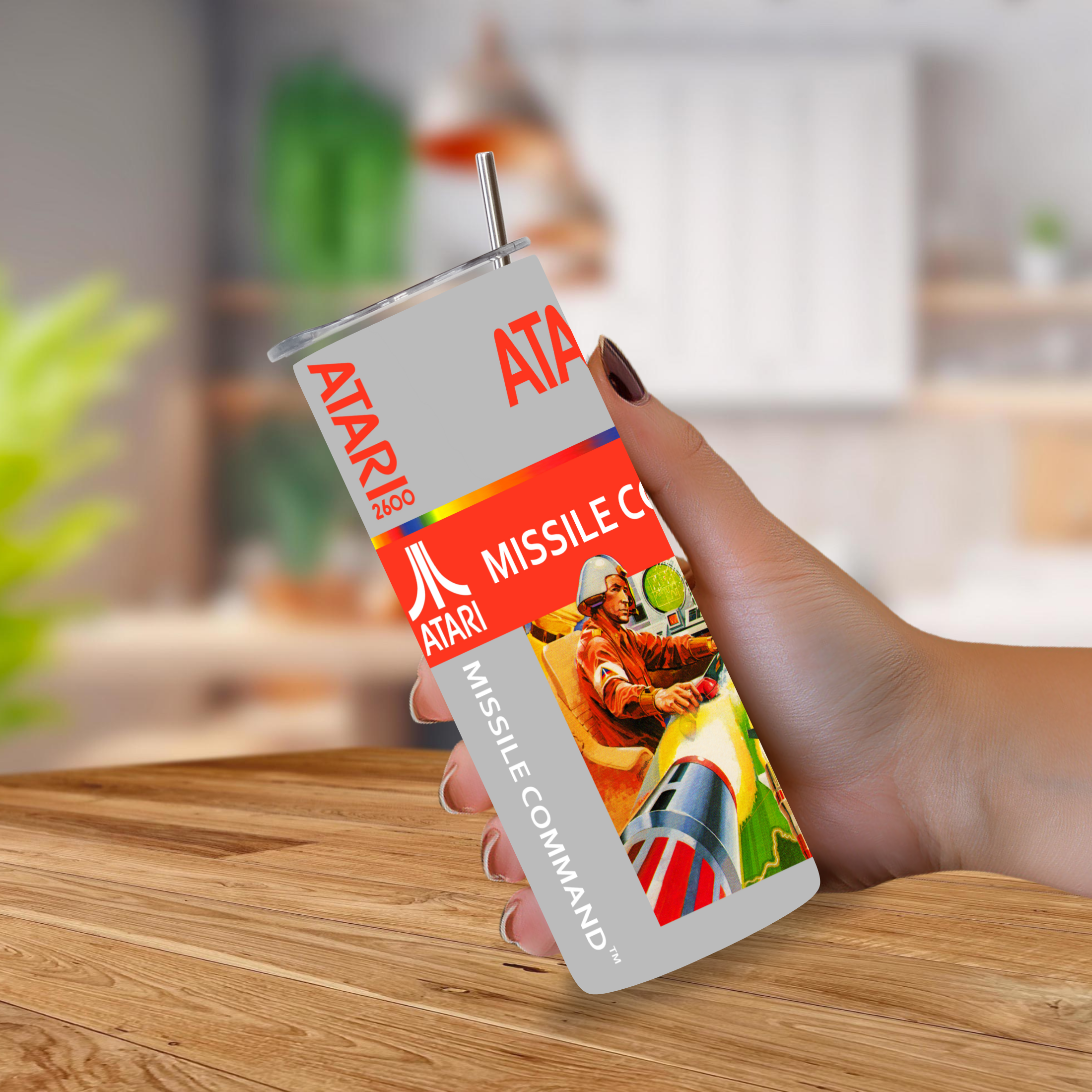 A hand holding a 20oz stainless steel tumbler with an Atari Missile Command straw, set against a blurred kitchen background.